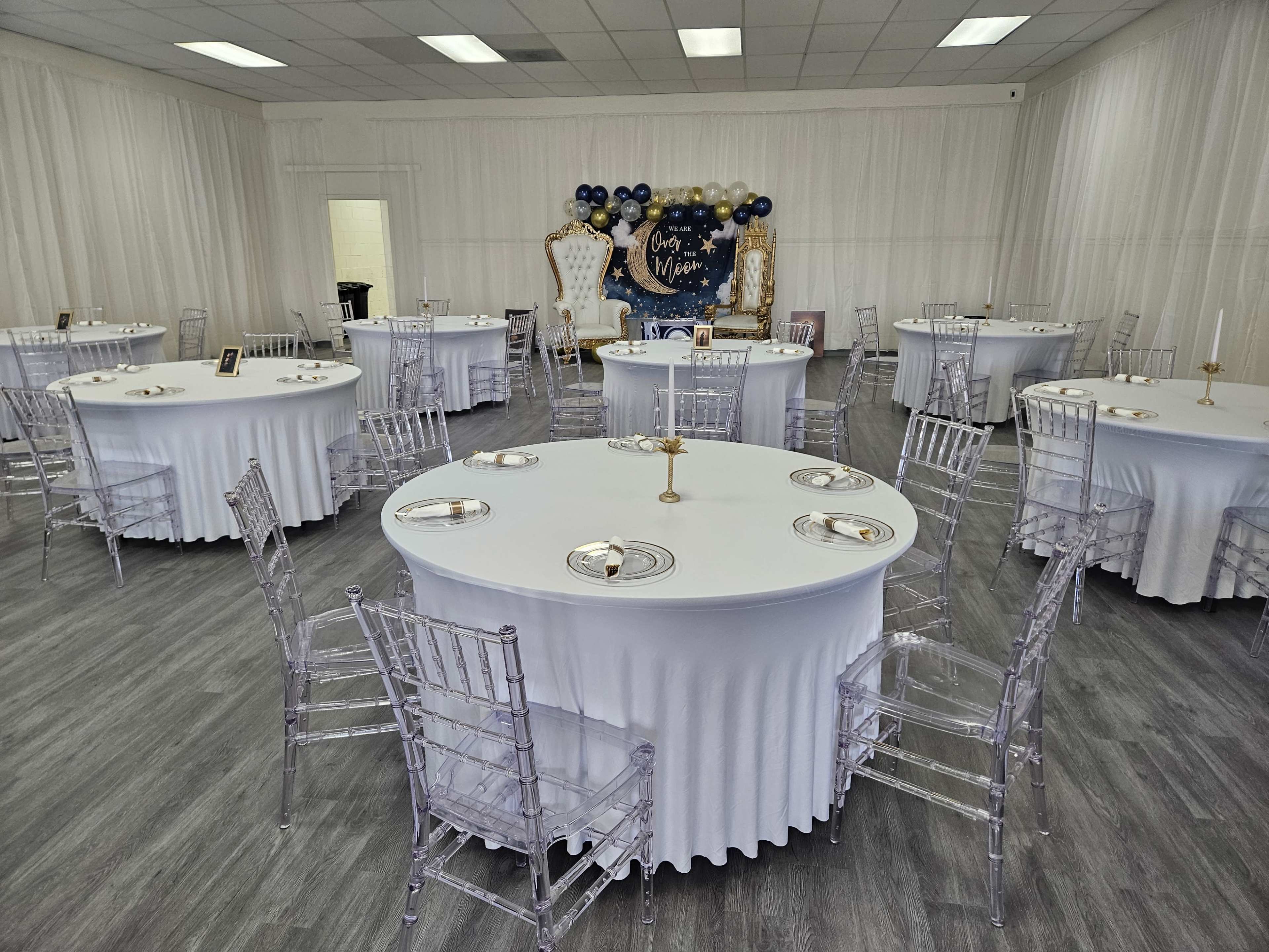 A spacious event room arranged with several round tables covered in white tablecloths and surrounded by clear acrylic chairs, set for a formal gathering.