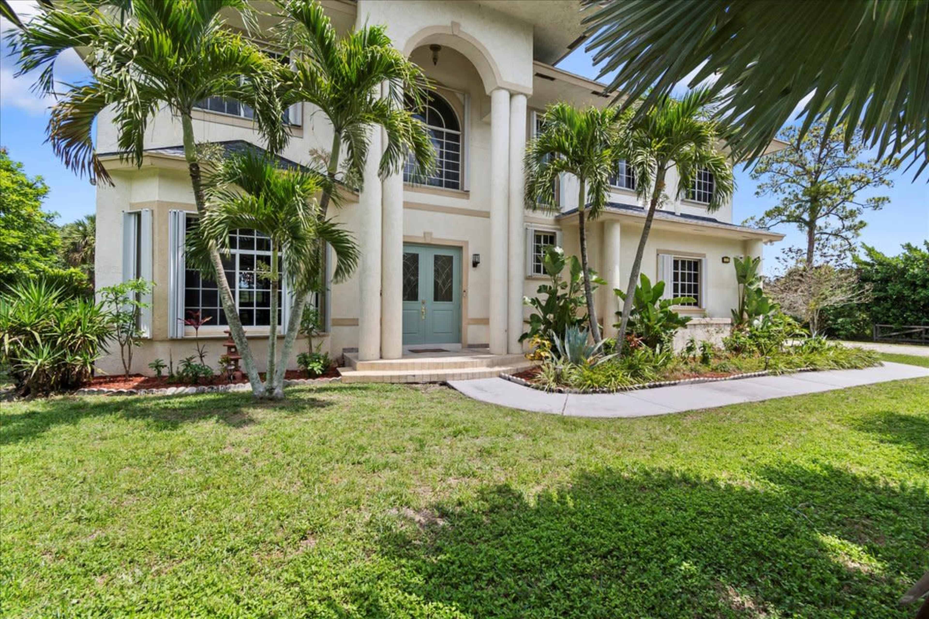 A two-story house with large windows and a charming porch is surrounded by palm trees and greenery.