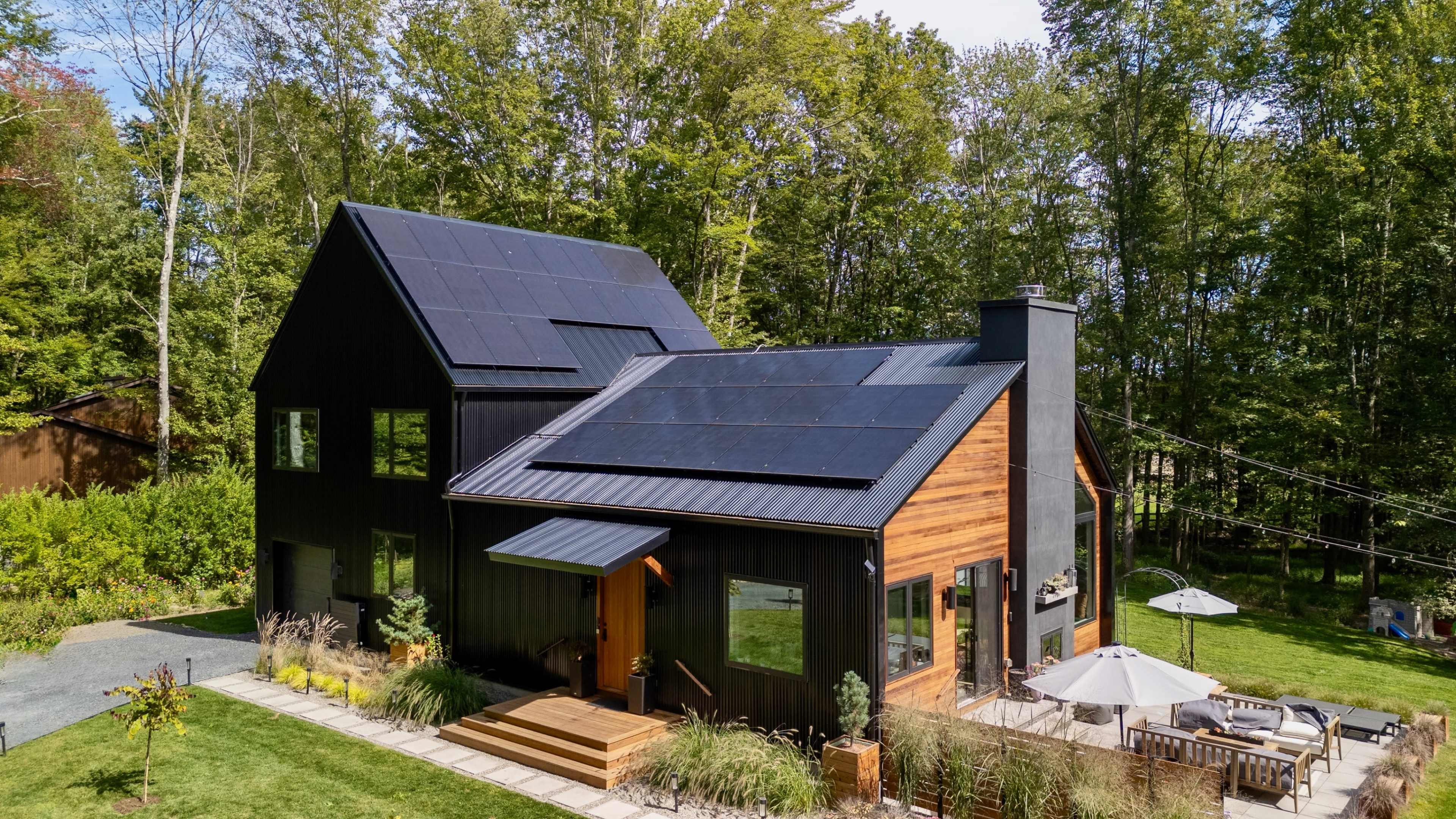 The image shows a modern black and wood house with solar panels on the roof, situated in a wooded area with a patio and outdoor furniture.