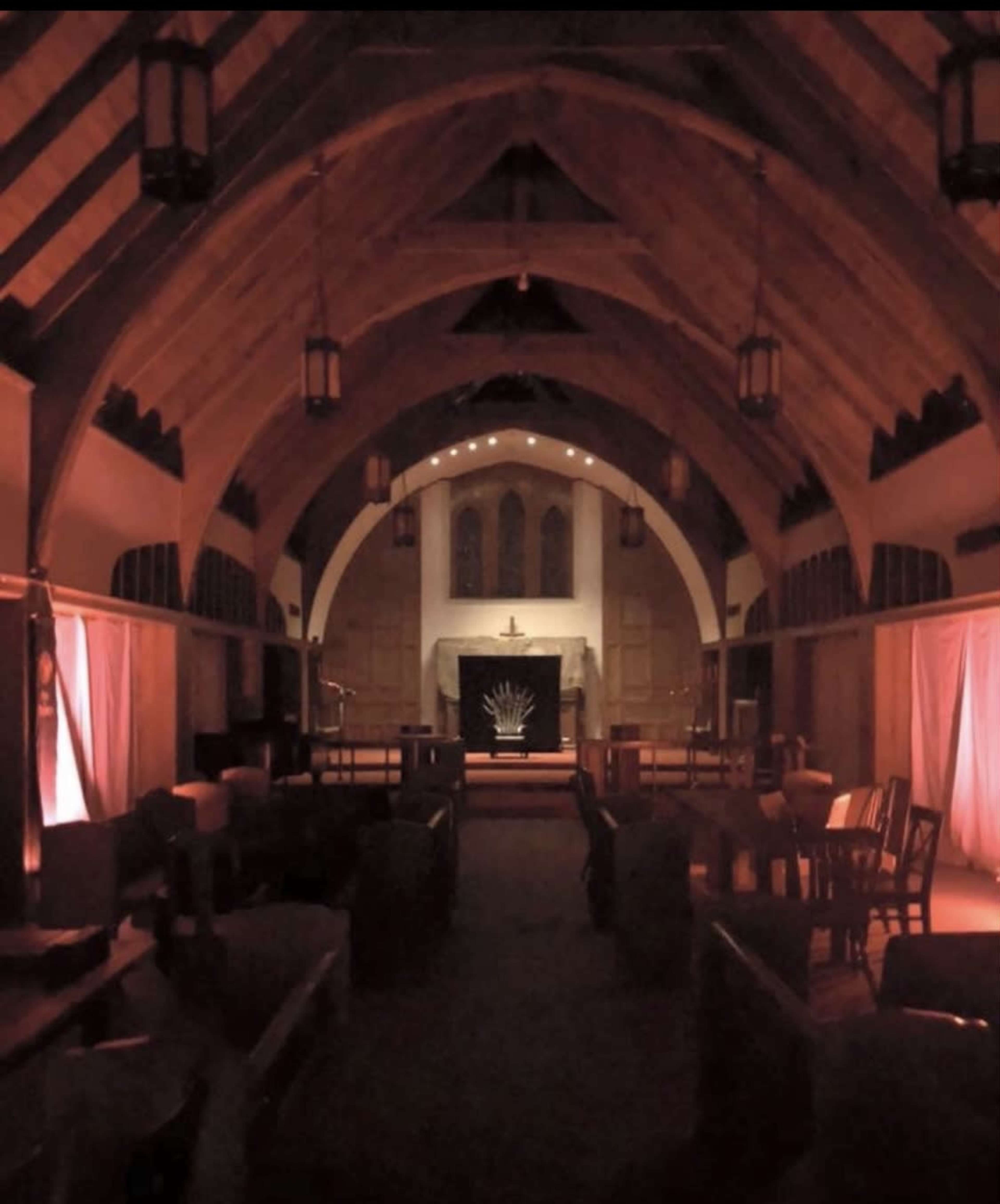The image shows a dimly lit interior of a large hall with wooden beams, a fireplace at the far end, and seating arranged throughout the space.