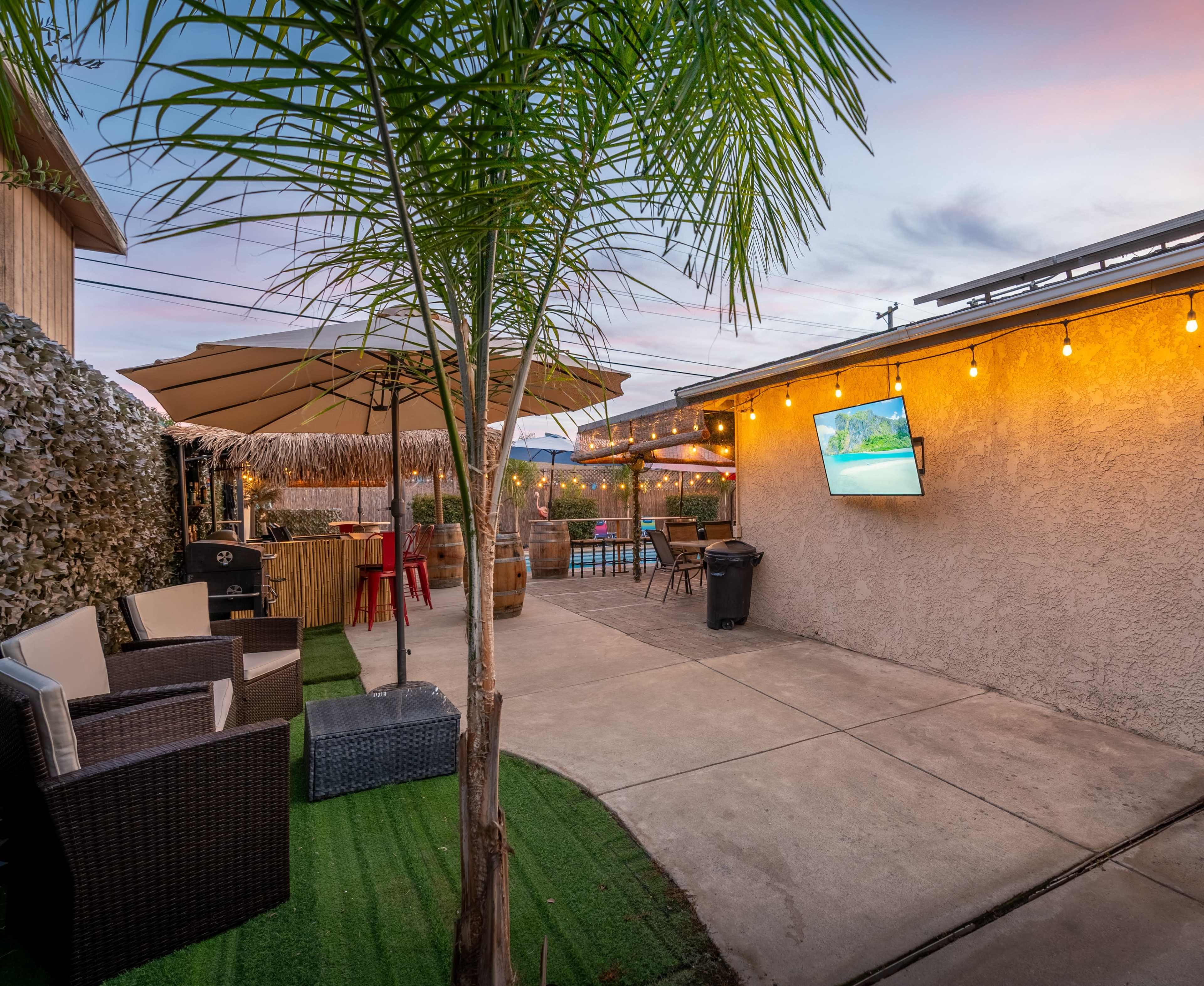 A backyard patio area featuring a seating arrangement, a large umbrella, string lights, and a mounted TV on the wall, all set against a backdrop of a swimming pool.