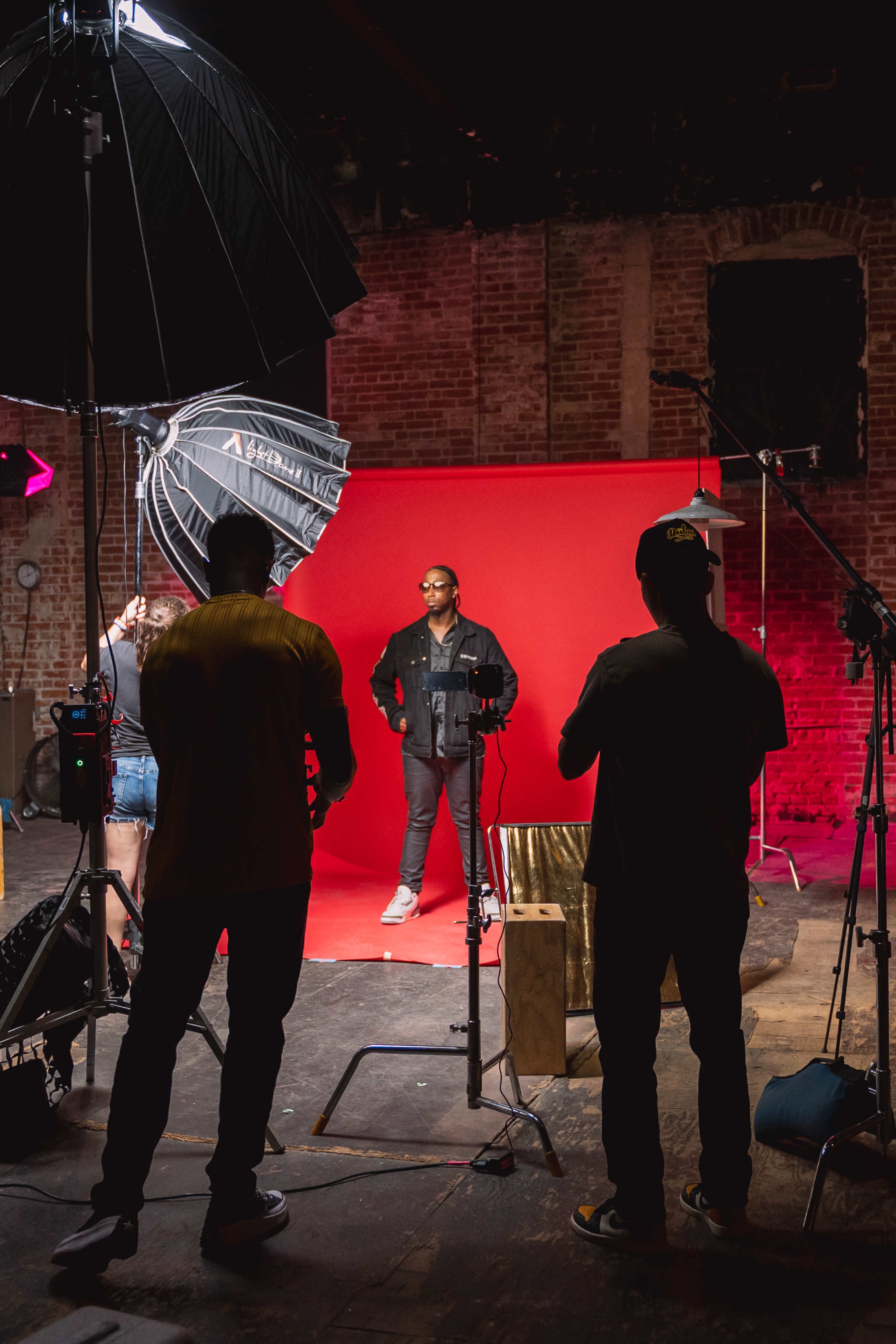 A photoshoot is in progress with a model posing against a bright red backdrop, while several photographers and assistants adjust lighting equipment.