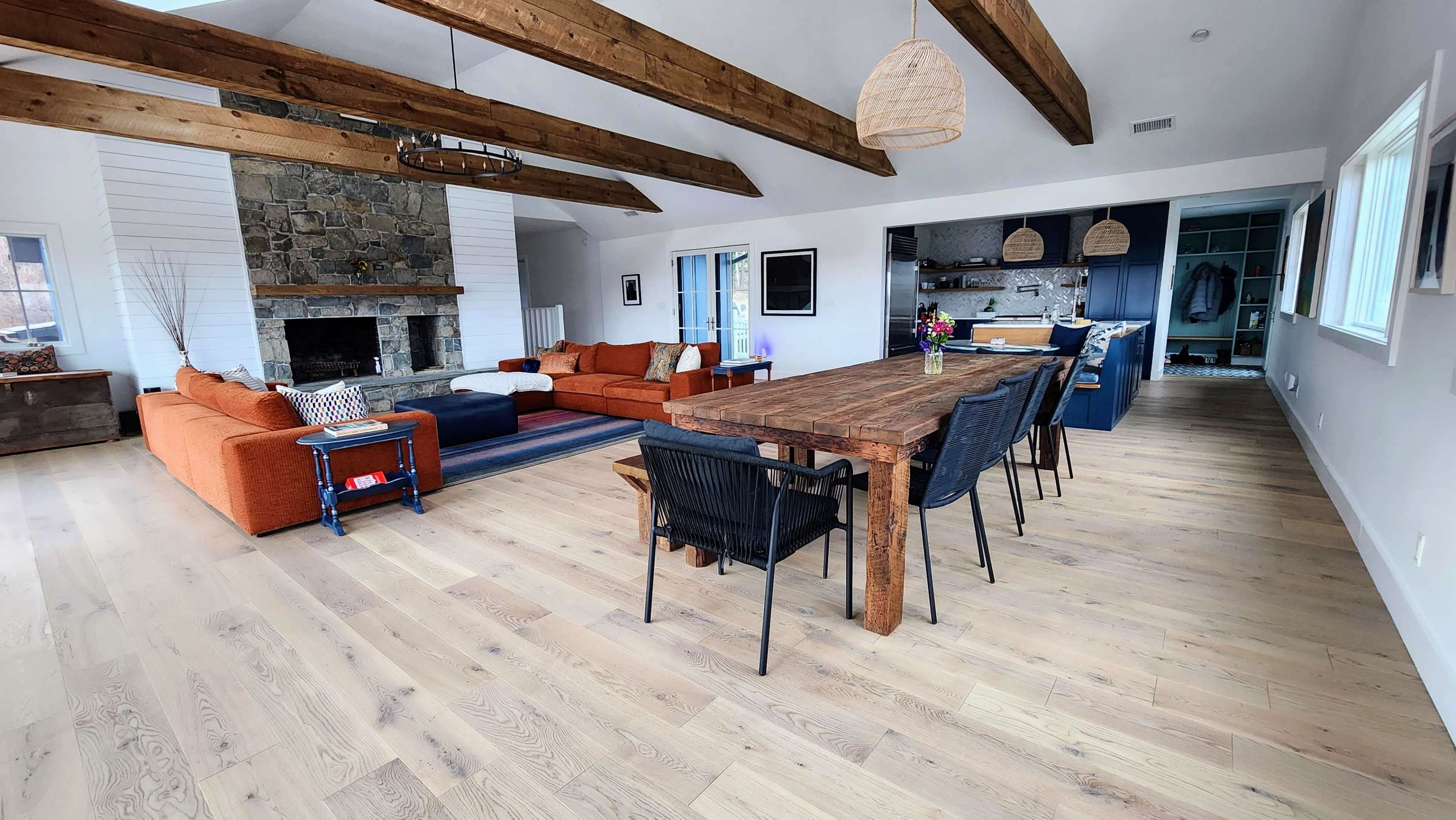 A spacious living and dining area with wooden beams, a stone fireplace, an orange sectional sofa, a long wooden dining table, and a well-lit kitchen in the background.