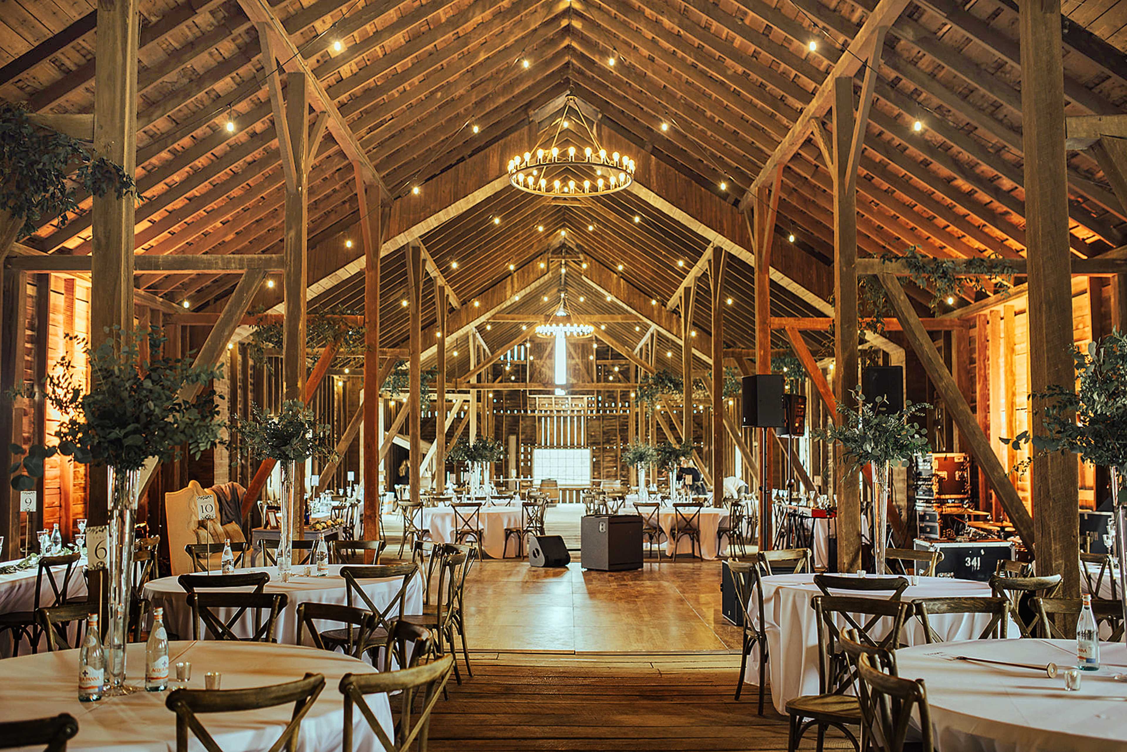 The interior of a spacious barn features round tables with white tablecloths, rustic wooden beams, and chandeliers hanging from the high ceiling.