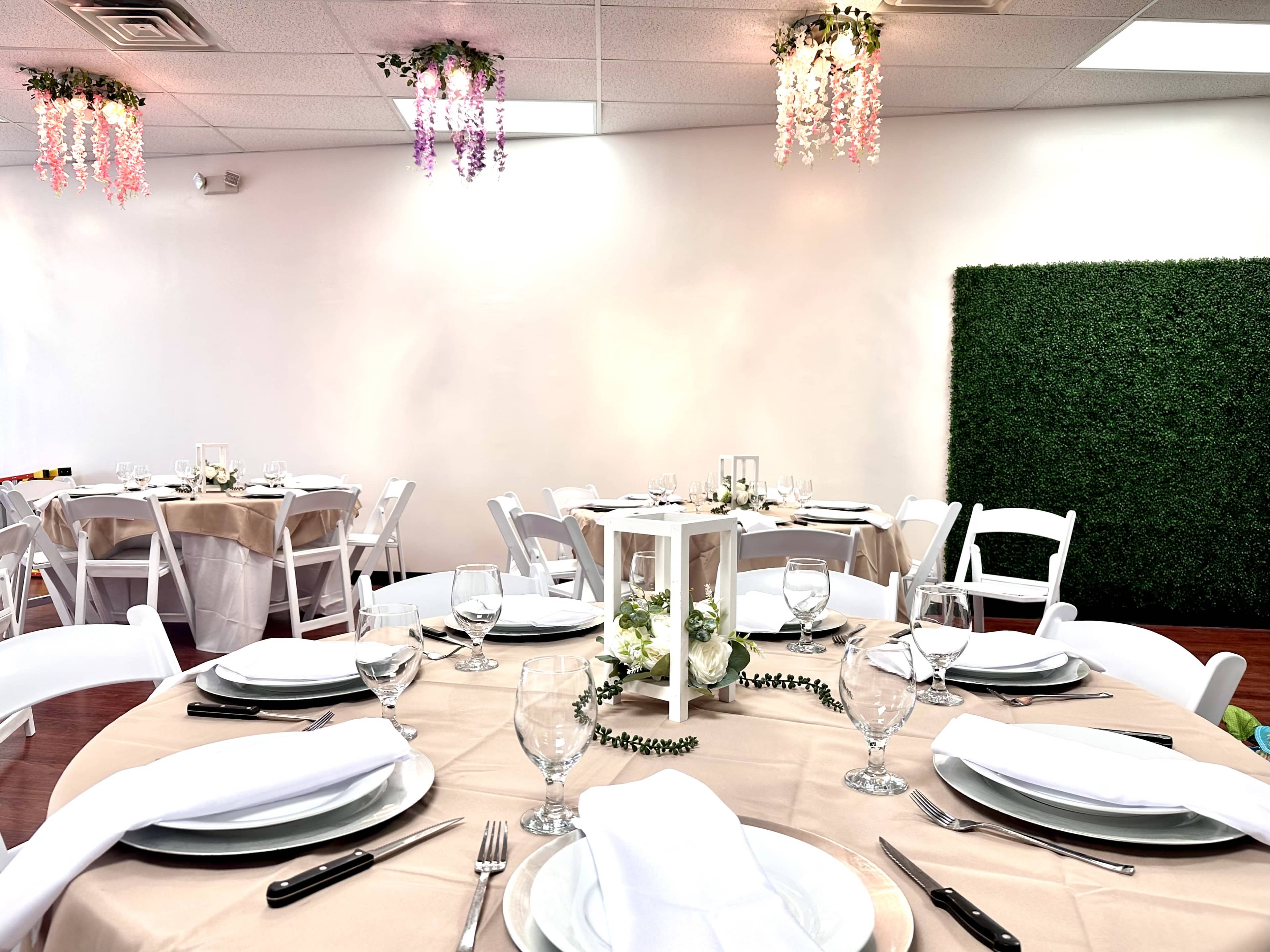A decorated dining area with tables set for an event, featuring white chairs, a beige tablecloth, and floral accents overhead.