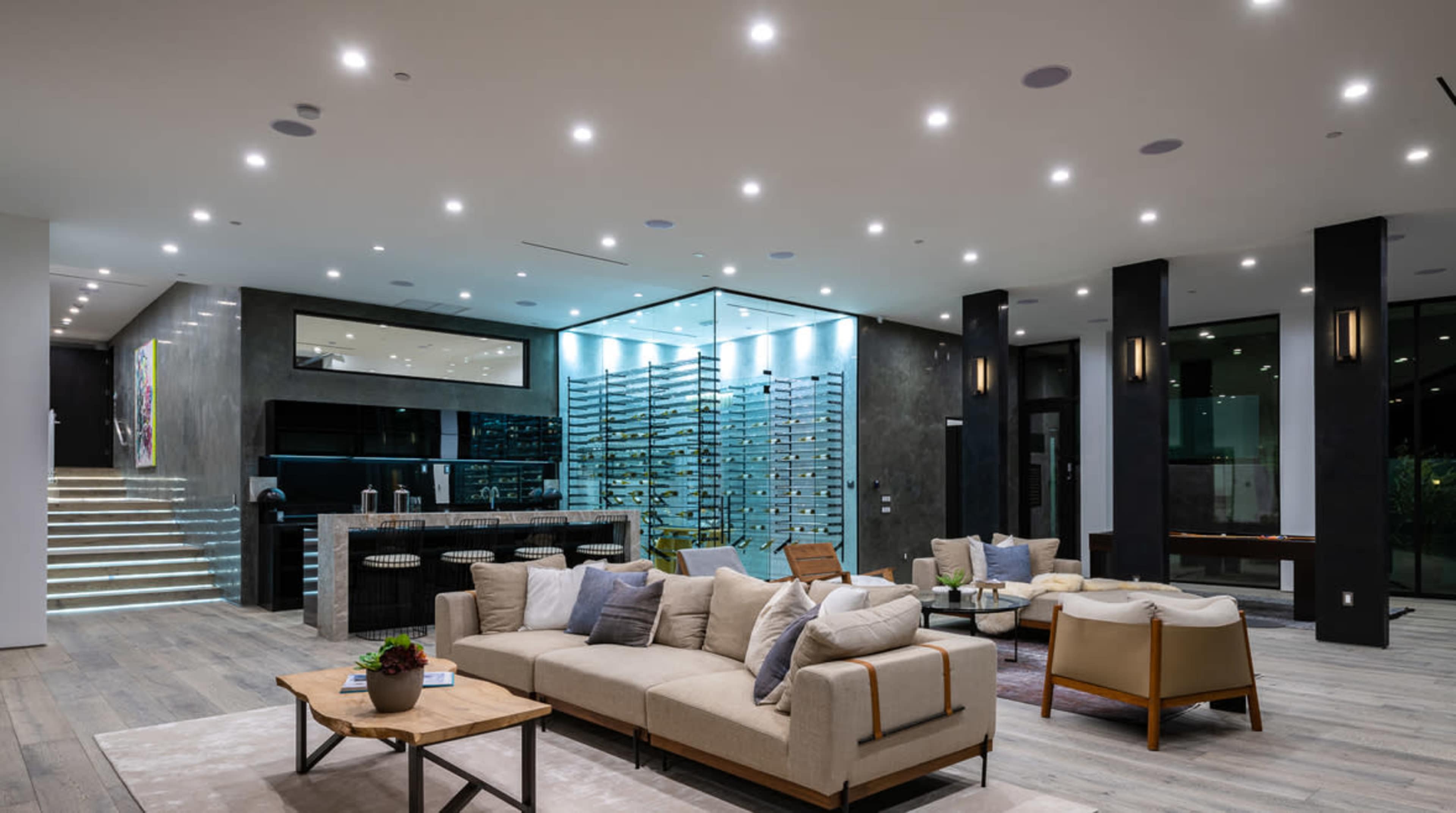 A modern living room features a spacious layout with a large beige sectional sofa, wooden coffee table, and a wine wall illuminated behind a sleek bar area.