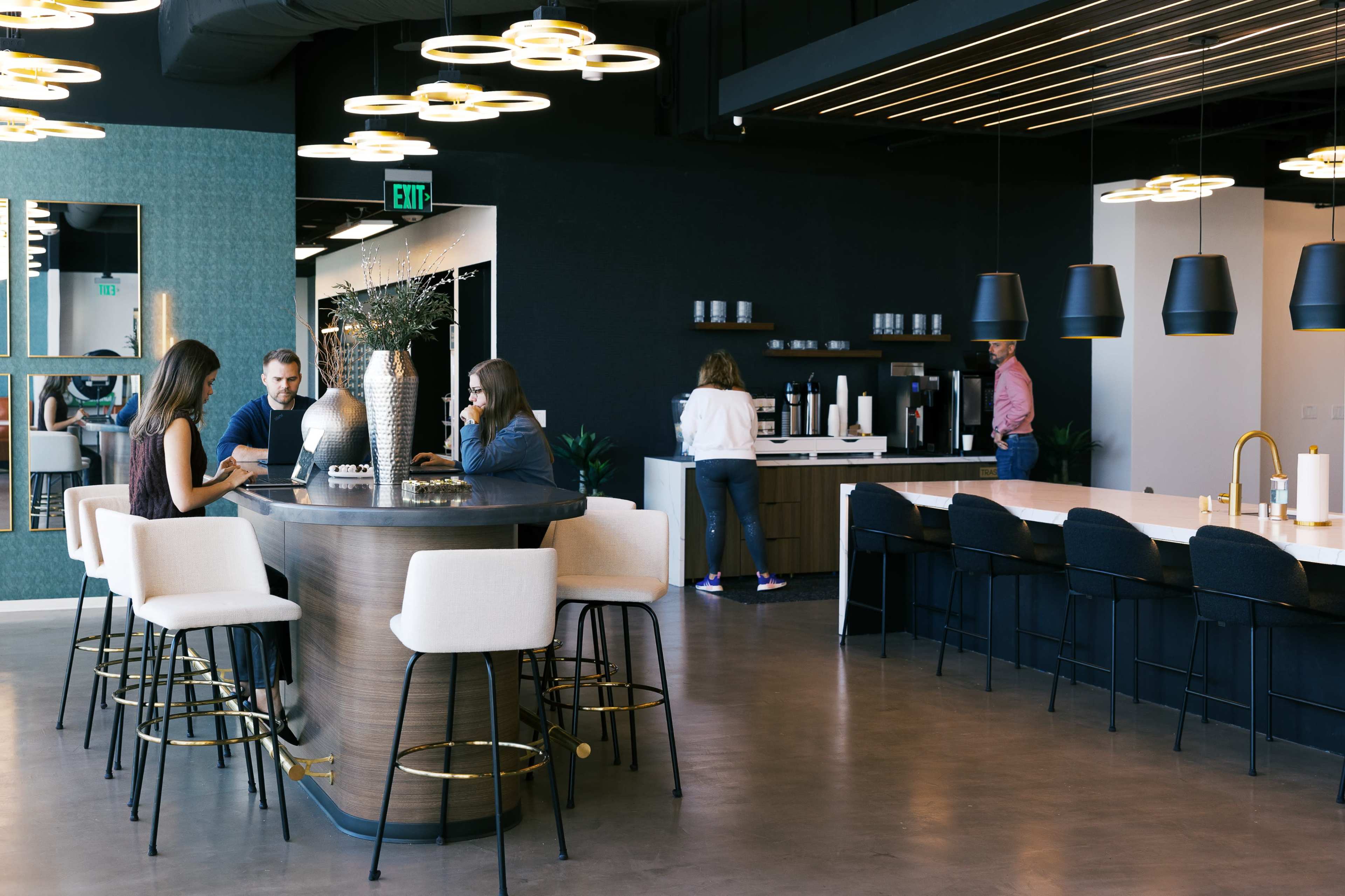 A modern office break room features a round table surrounded by bar stools, with several individuals engaging in conversation, while a coffee station is visible in the background.