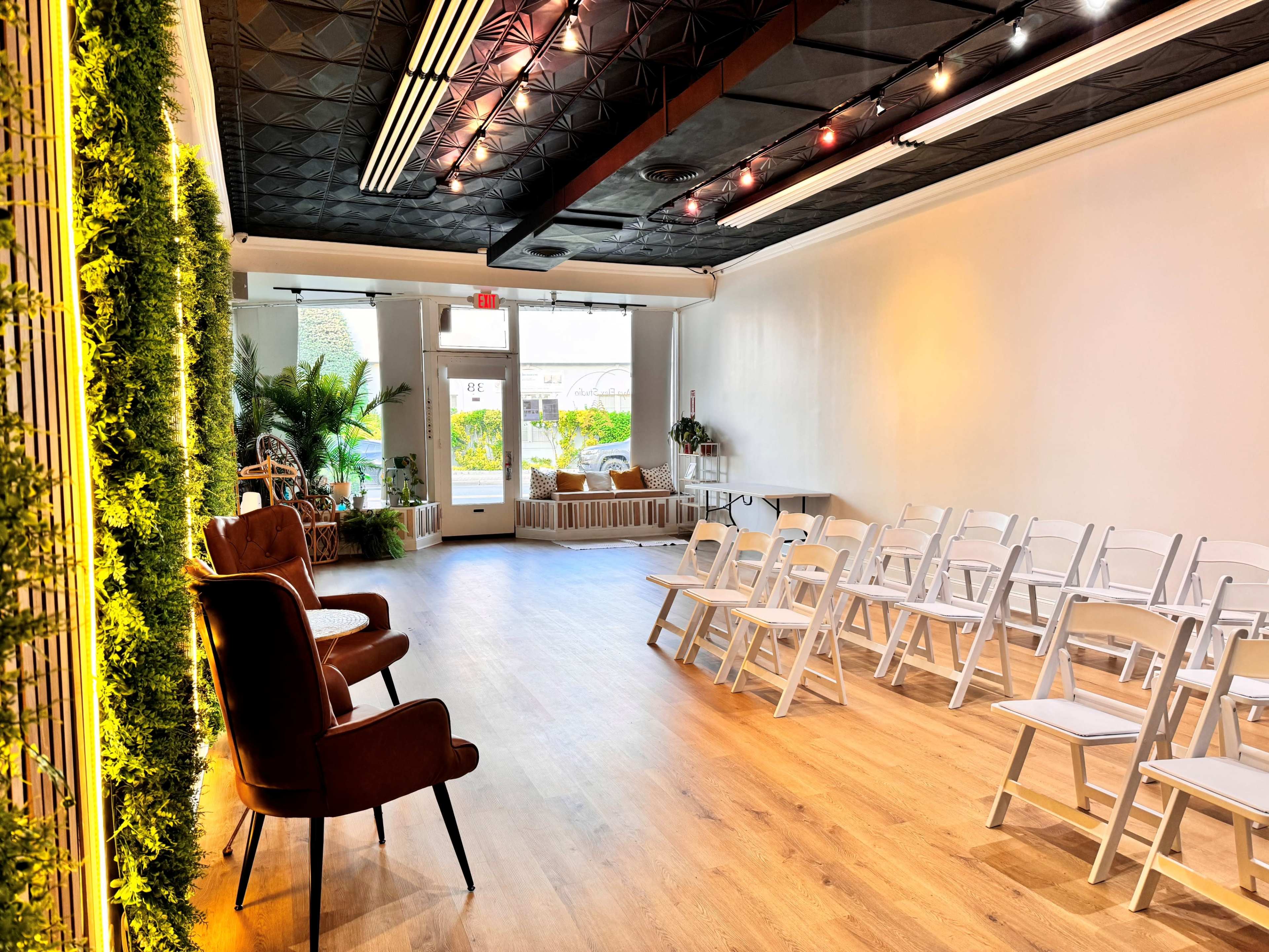 A spacious room features a seating arrangement of white folding chairs facing a blank wall, with two brown armchairs on one side and plants along the back.