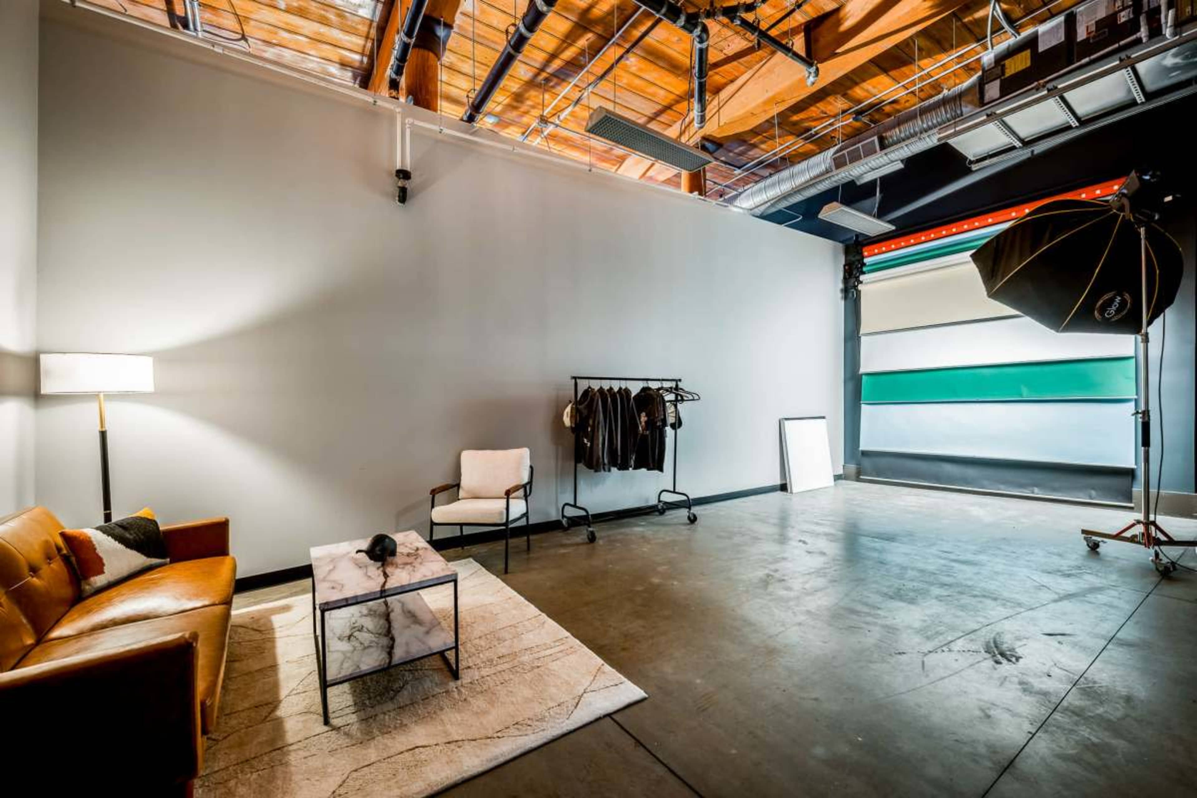 The image shows a spacious studio with a brown leather sofa, a small coffee table, a clothing rack with black garments, and large rolls of backdrop paper near an open garage door.