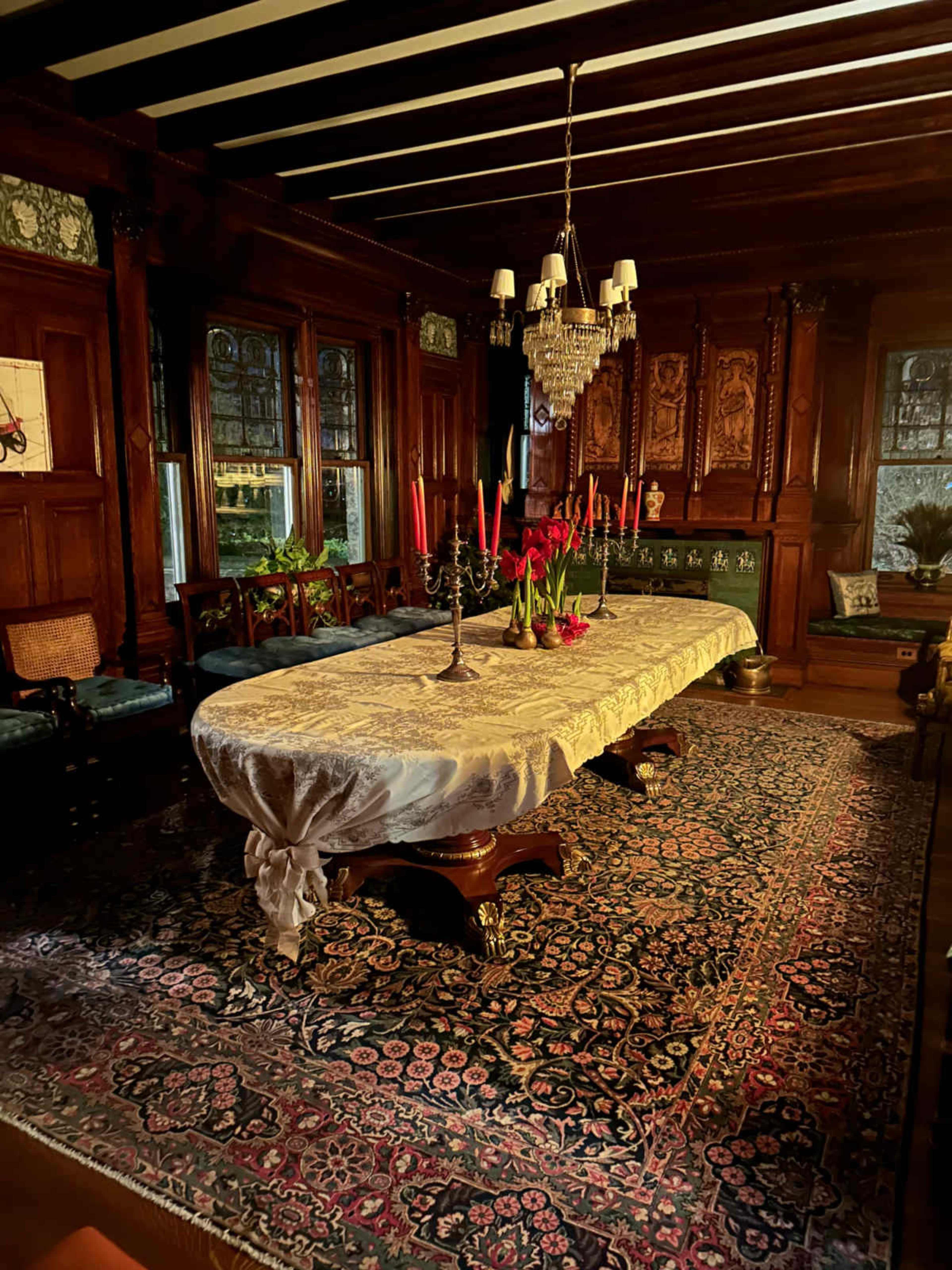 The image shows a large dining table covered with a tablecloth, surrounded by wooden chairs, in a richly decorated room featuring dark wood paneling and an ornate carpet.