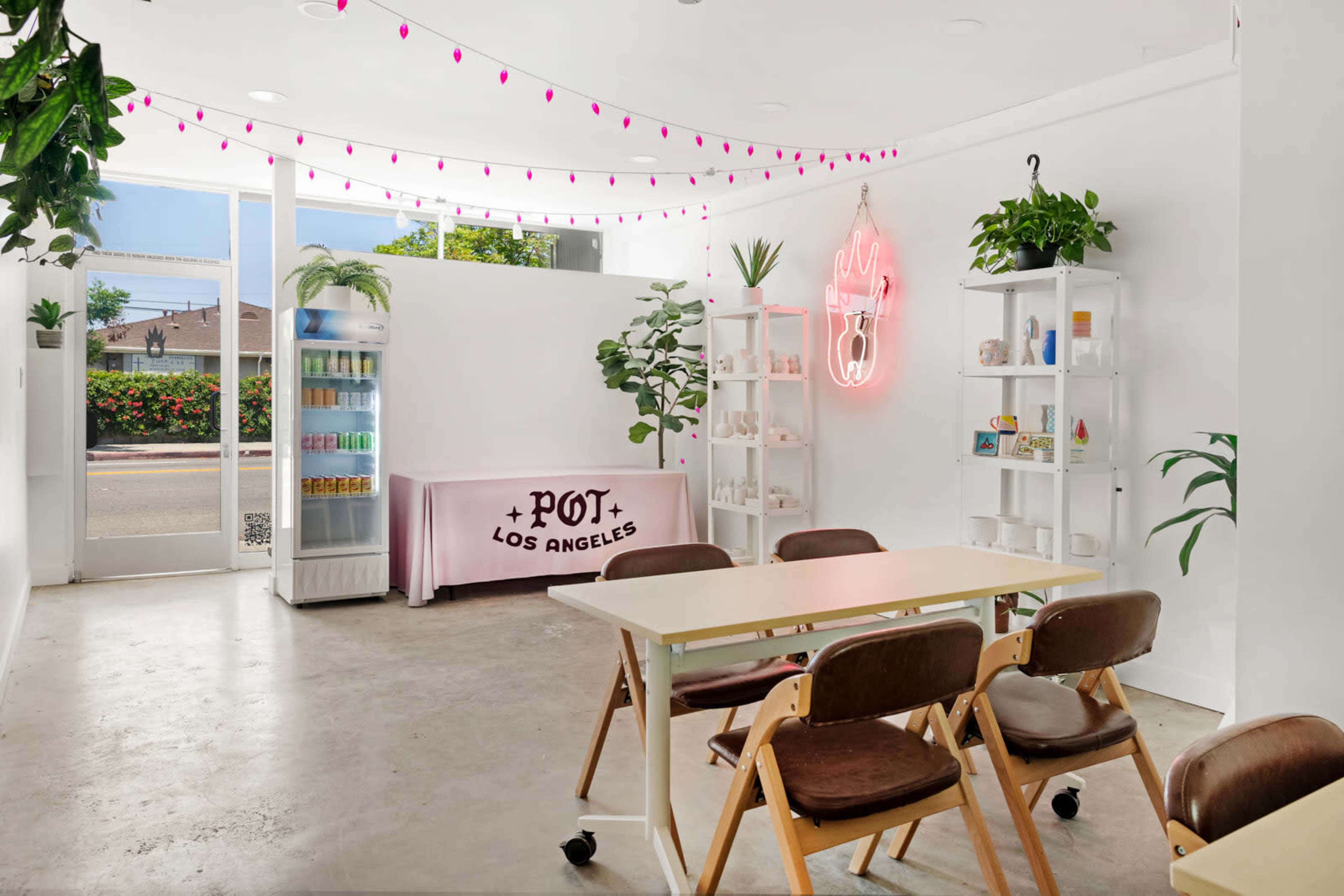 The interior of a bright, minimalist room features a large window, a table with chairs, shelves filled with items, and a neon sign.