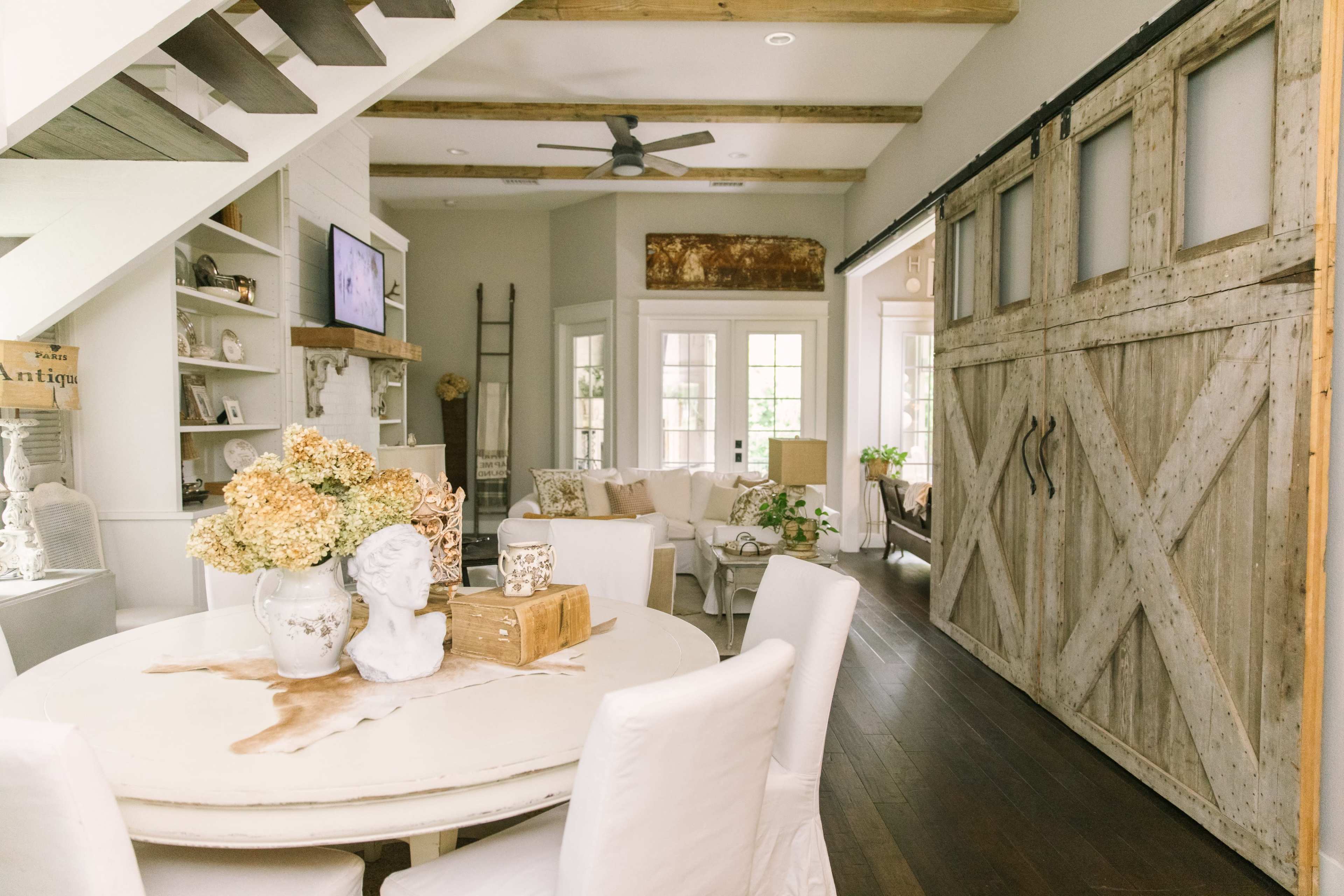 The image shows a cozy interior space featuring a round dining table with white chairs, a rustic sliding barn door, and a view of a living area with large windows.
