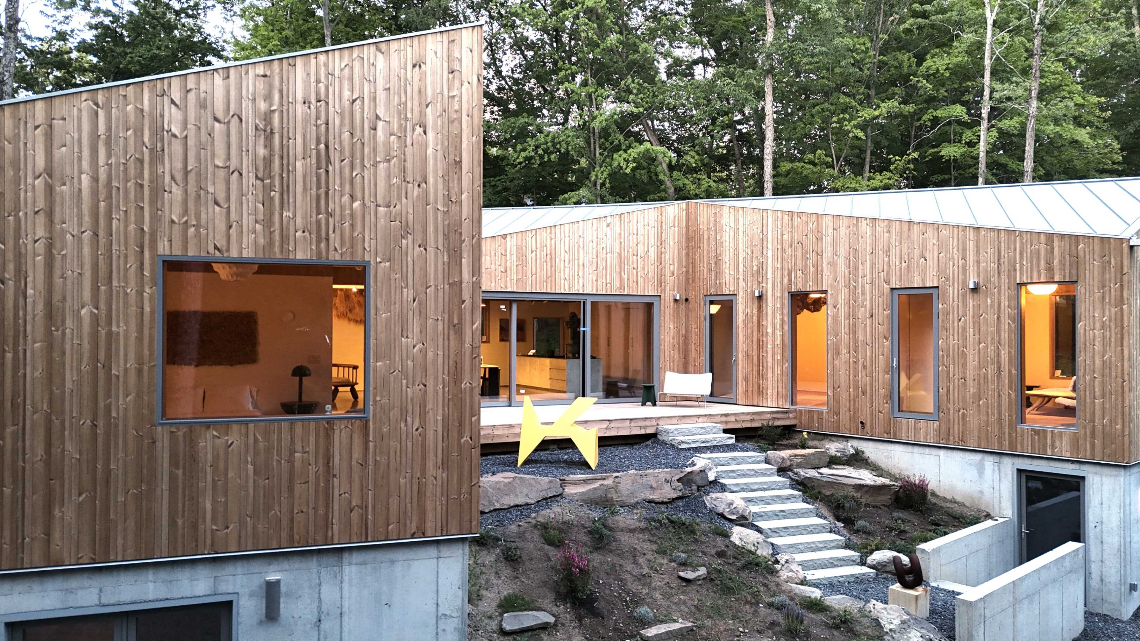 The image shows a modern wooden house with large windows, featuring a stone pathway leading to an outdoor area with a yellow star sculpture.