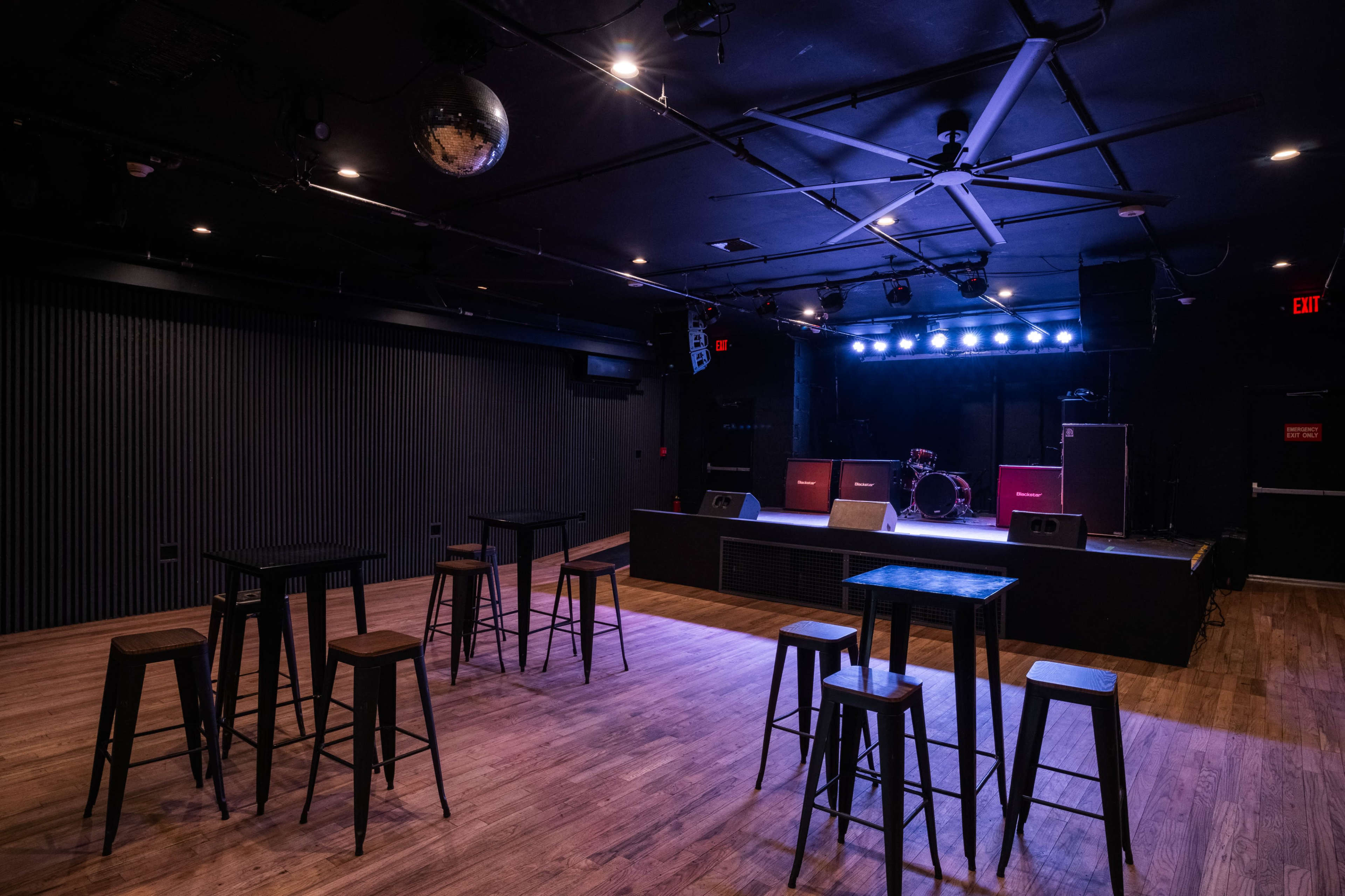 The image shows a dimly lit music venue with a small stage, equipped with amplifiers and a drum set, surrounded by several tall tables and stools on a wooden floor.