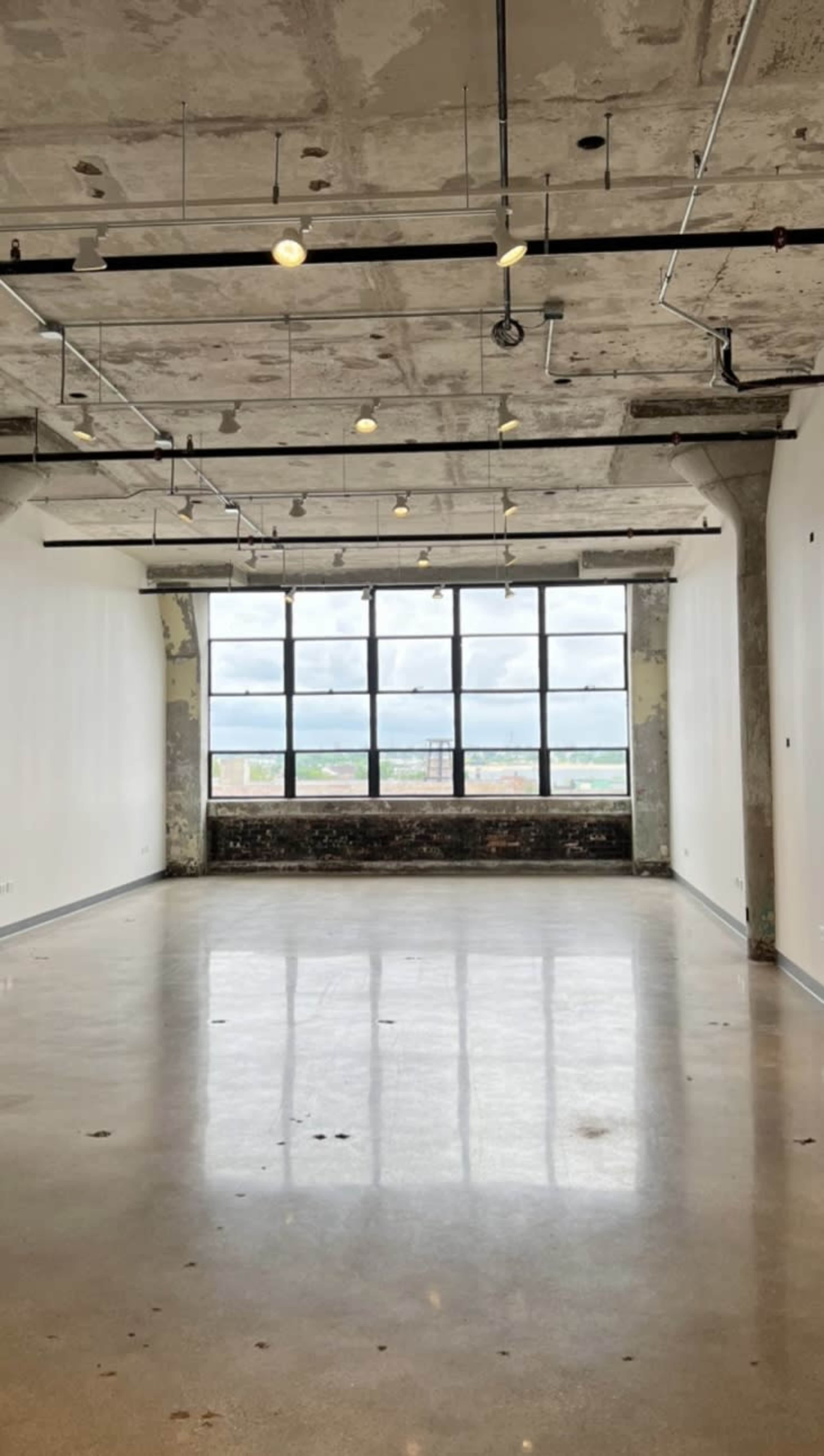 The image shows a spacious, industrial-style room with polished concrete flooring, exposed ceiling fixtures, and a large window revealing a view outside.