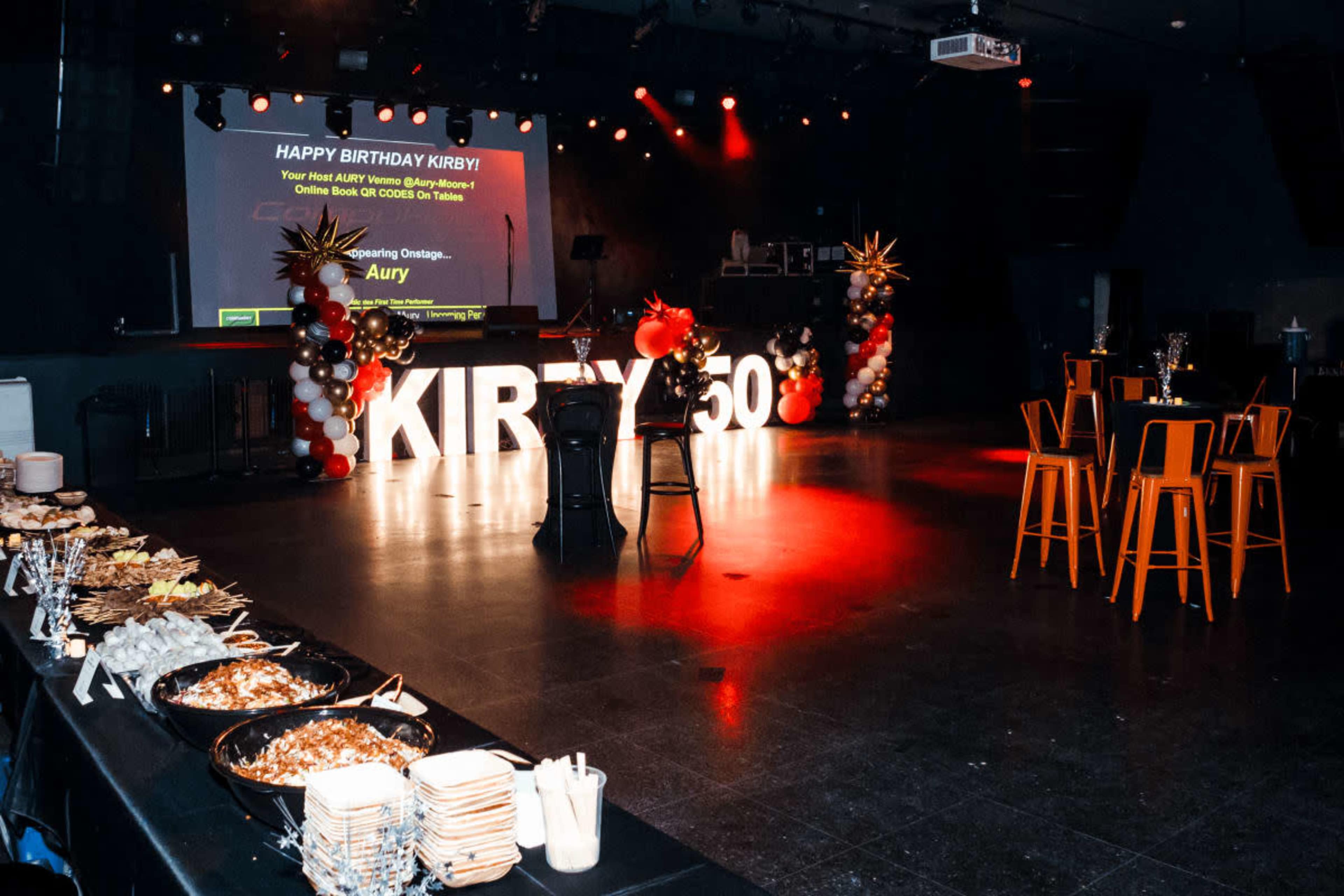 The scene features a birthday party setup with large illuminated letters spelling "KIRBY 50," a decorated stage in the background, and tables arranged with food and balloons.