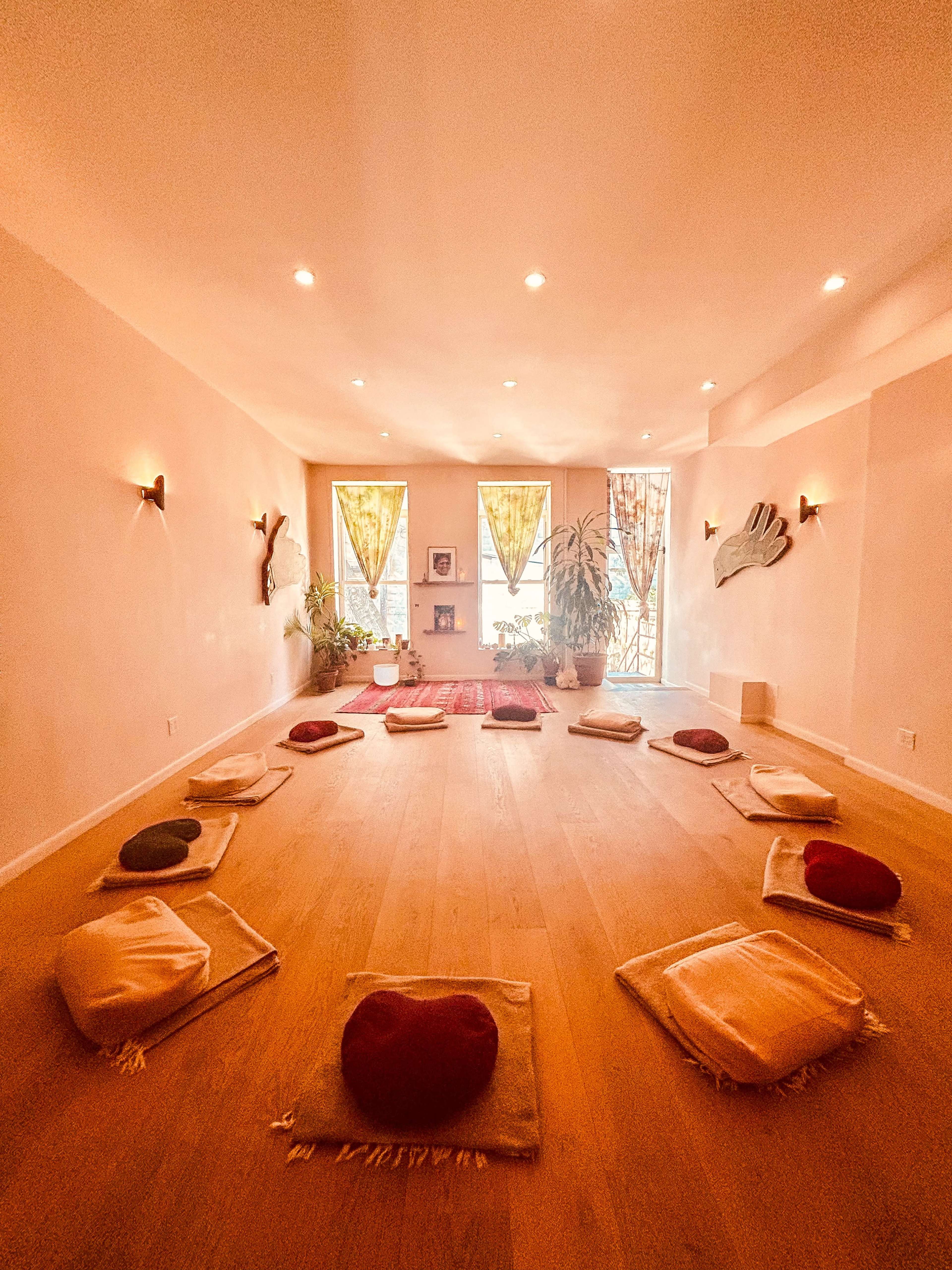 The image shows a meditation room with circular seating arrangements of cushions on the wooden floor, surrounded by plants and softly lit by natural light from large windows.