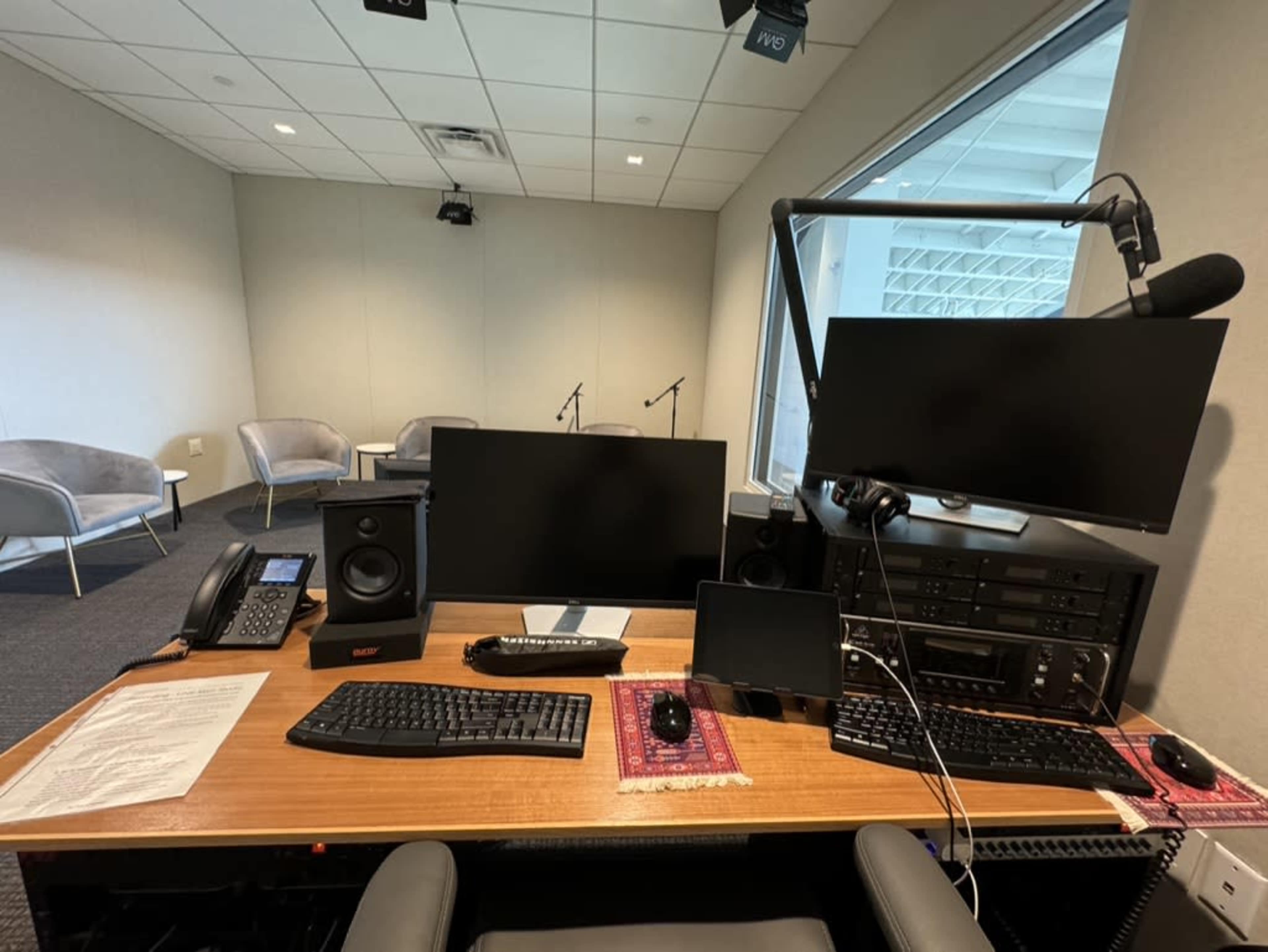 The image shows a radio or recording studio setup featuring a desk with a computer monitor, audio equipment, a keyboard, a mouse, and a phone, surrounded by empty seating areas.