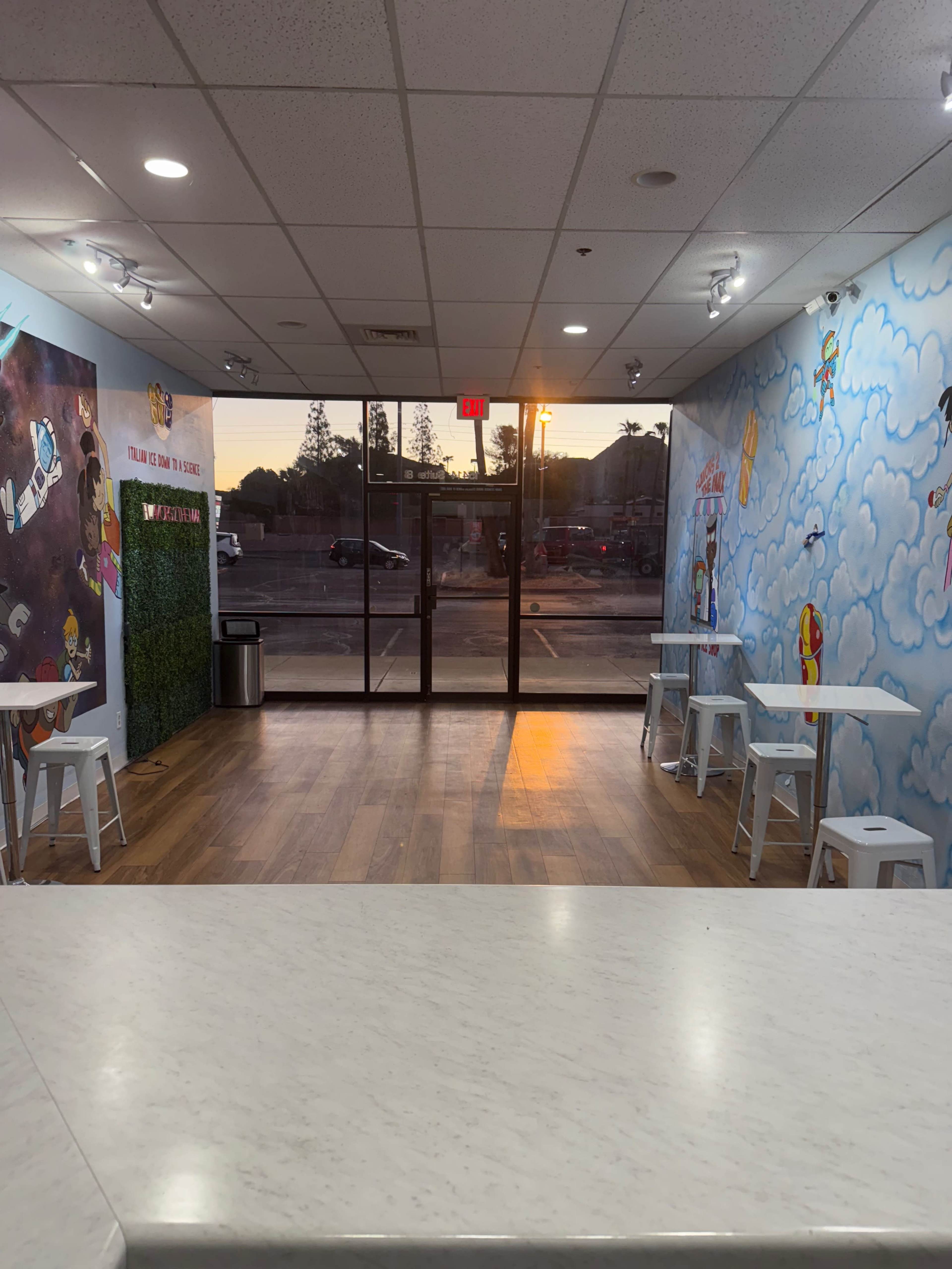 The image shows a brightly lit modern cafe interior with colorful wall murals, small white tables, and a glass entrance facing a parking area during sunset.