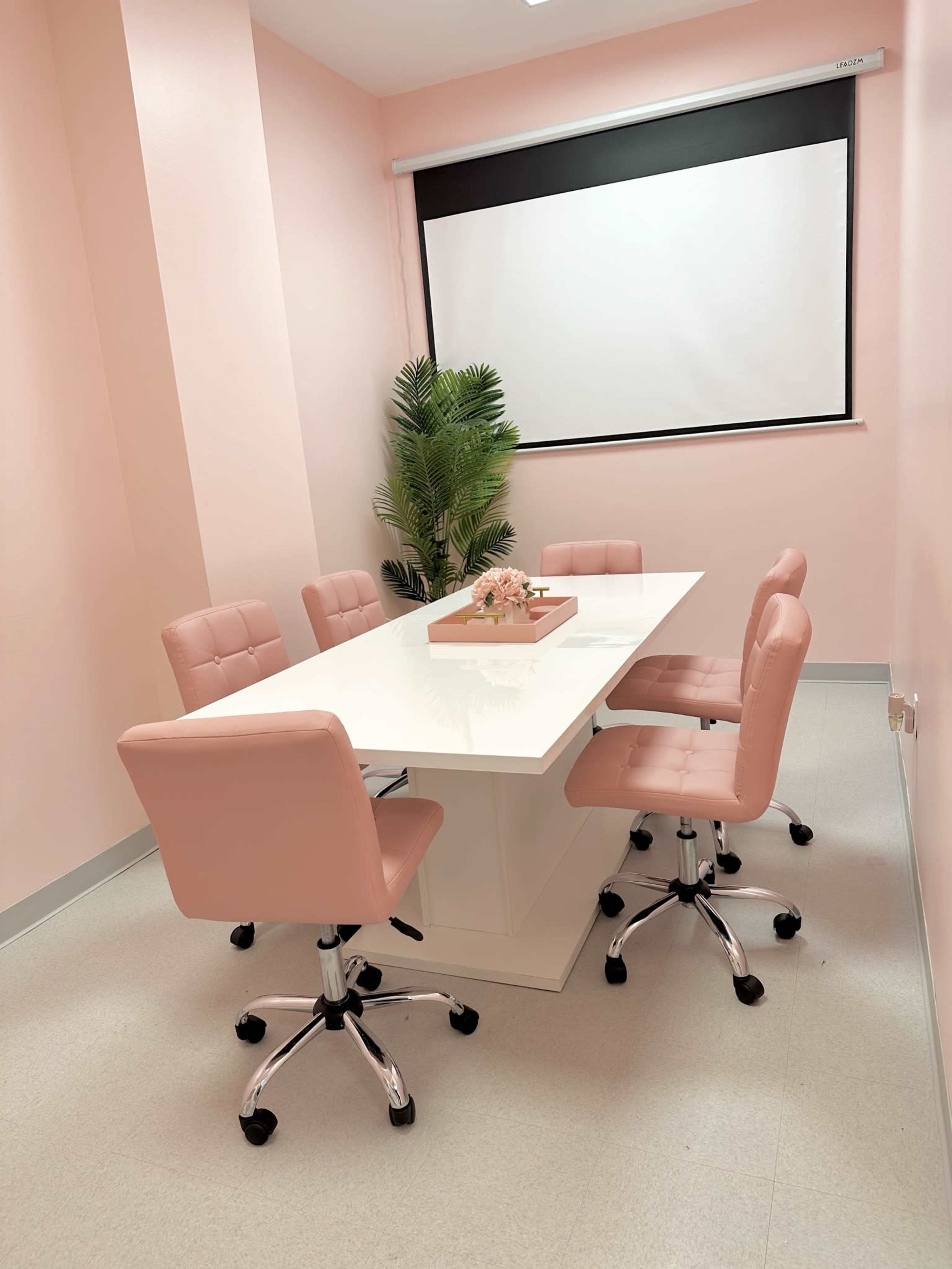 A conference room features a white table surrounded by pink chairs and a small decorative plant, with a projection screen at the front.