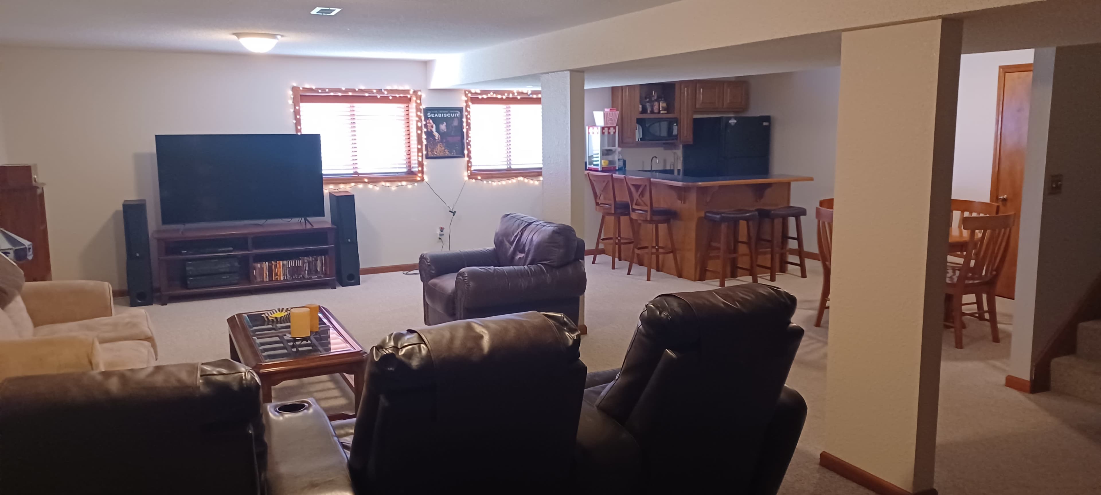 The image shows a finished basement with a seating area featuring two brown leather recliners, a television, and a bar with stools in the background.
