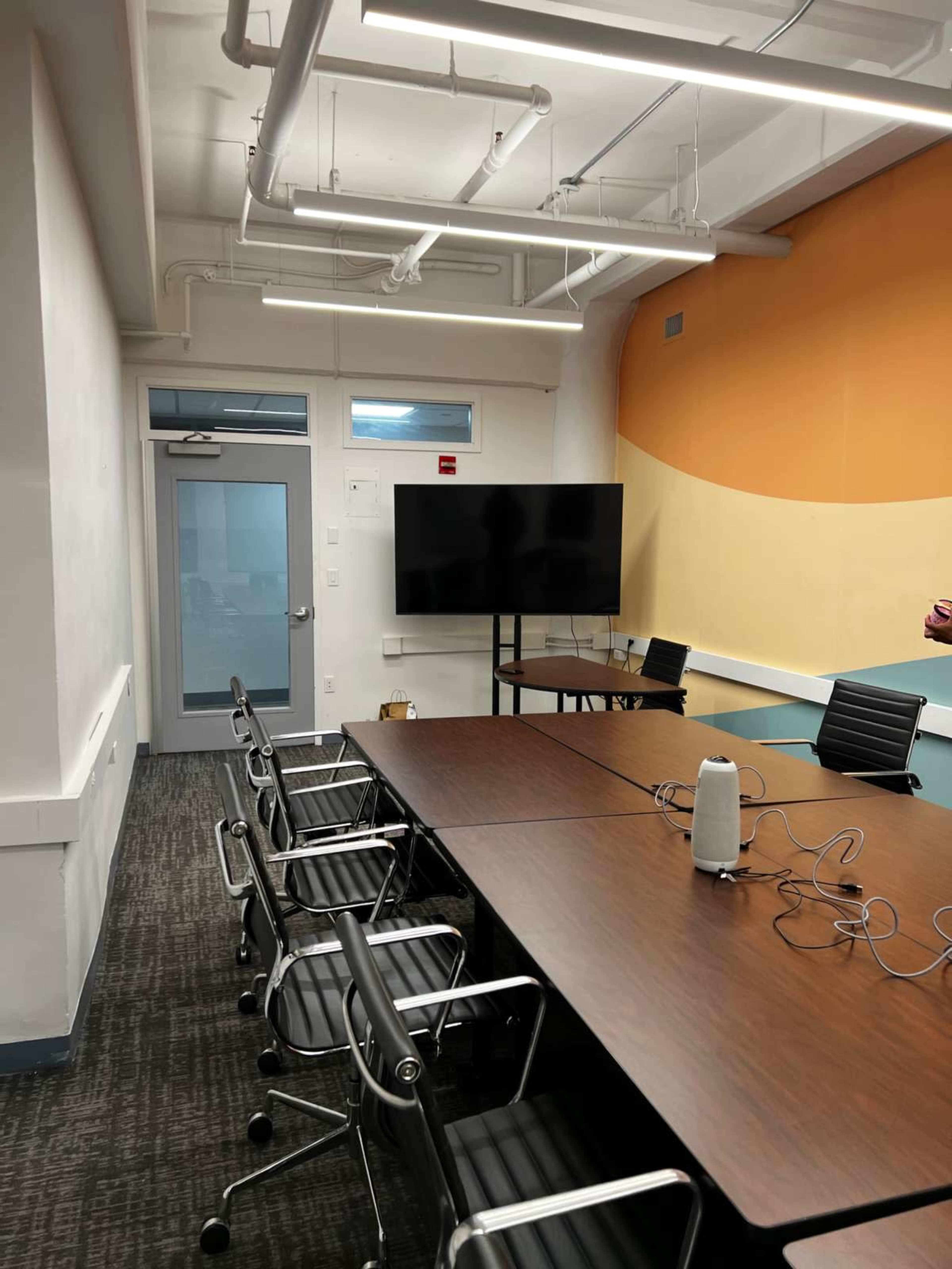 A modern conference room features a long wooden table, several black chairs, a large wall-mounted screen, and a door leading to an adjacent area.