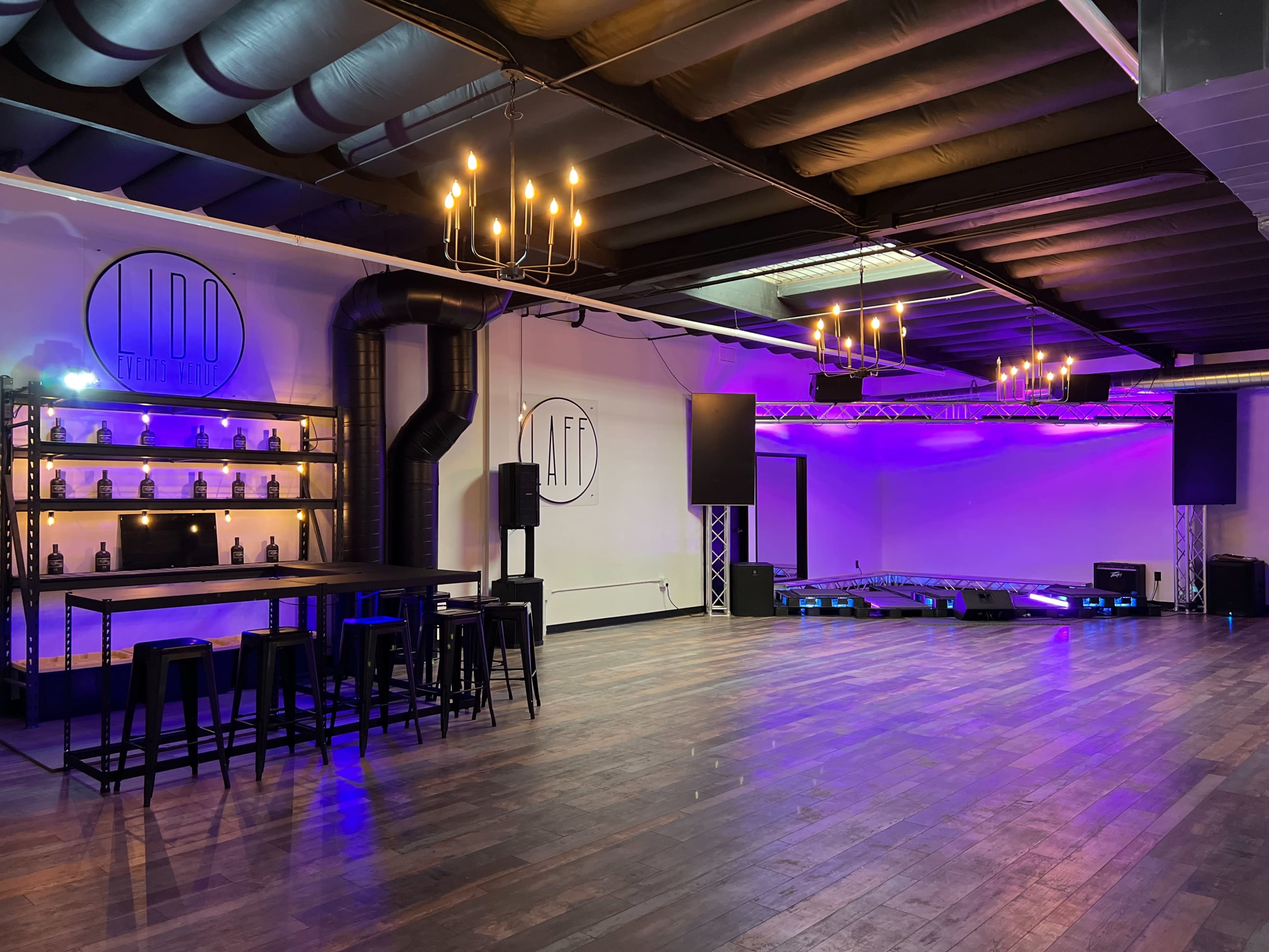 A modern event space with a wooden floor, shelving displaying bottles, and purple lighting accentuated by chandeliers.