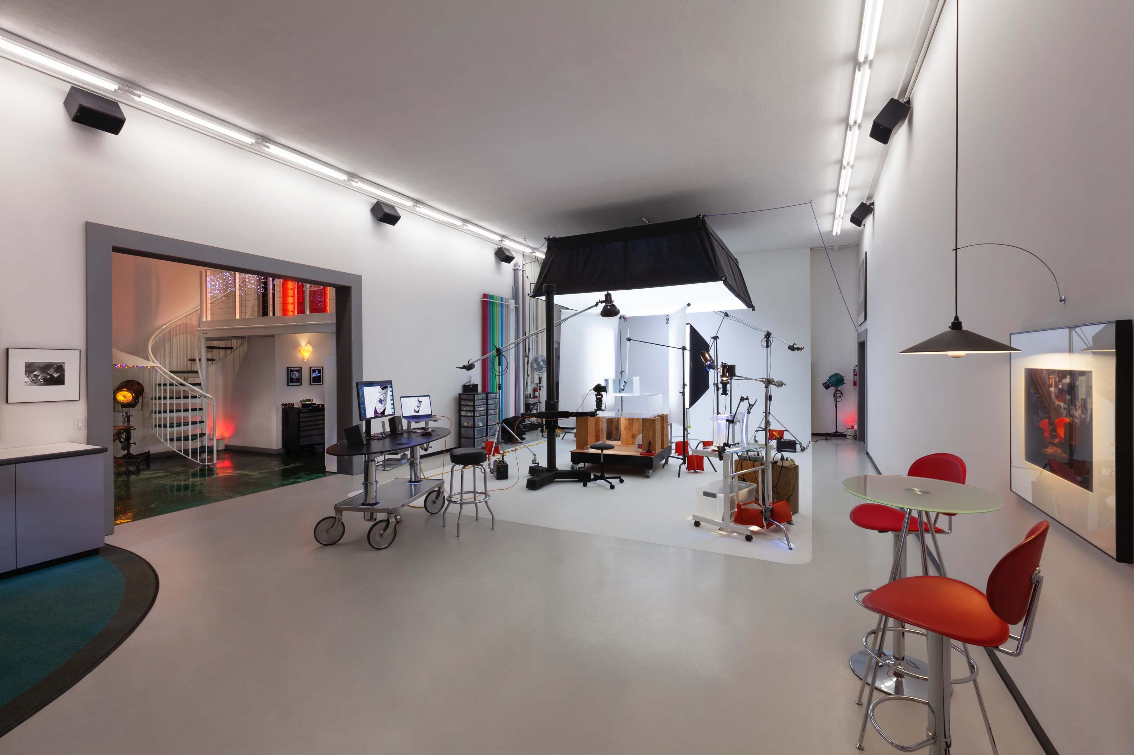 A spacious, modern studio with a variety of equipment, including a large backdrop and lighting set-up, alongside a worktable and seating areas.