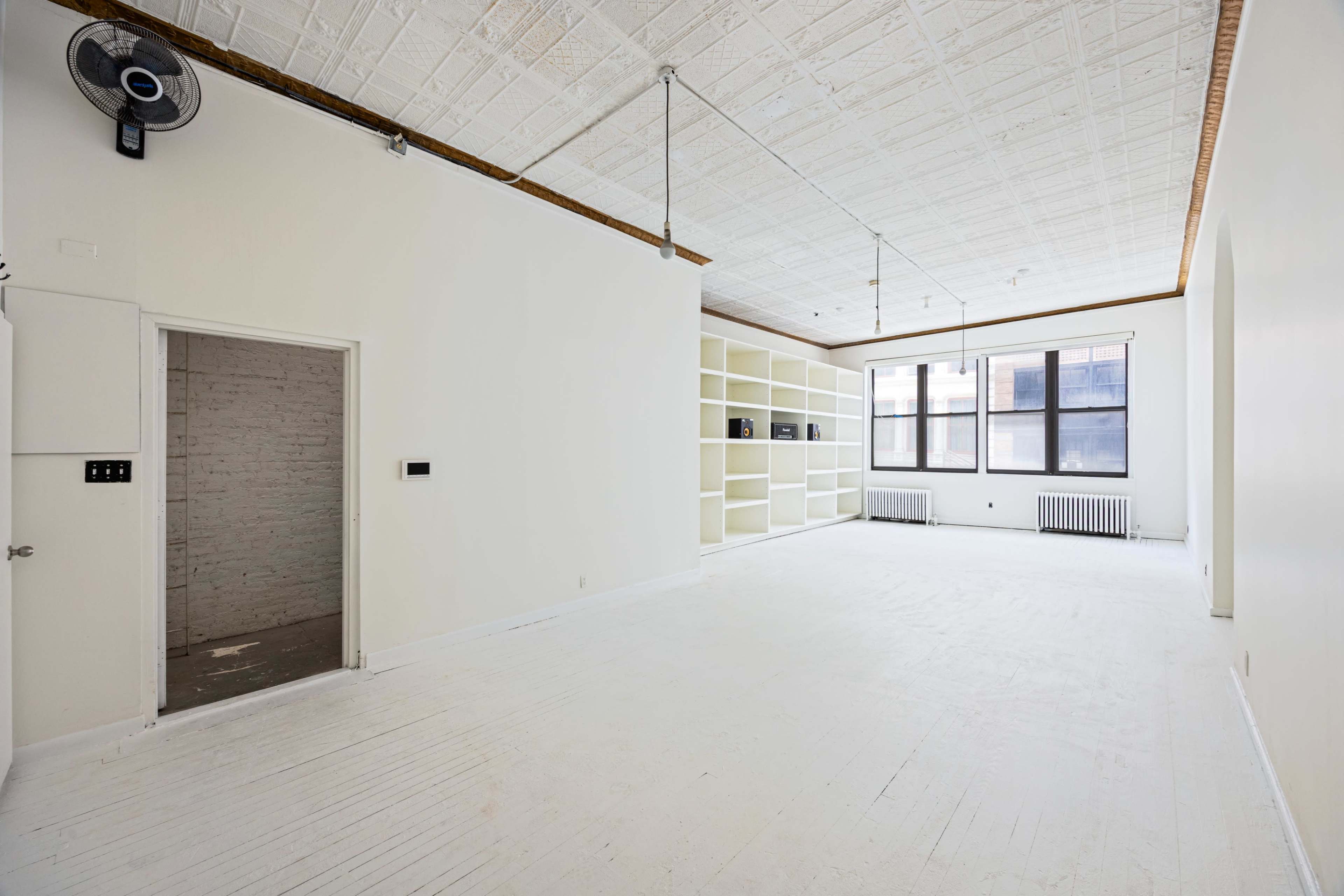 The image shows a spacious, empty room with white walls, a ceiling fan, and large windows, featuring built-in shelves on one side.