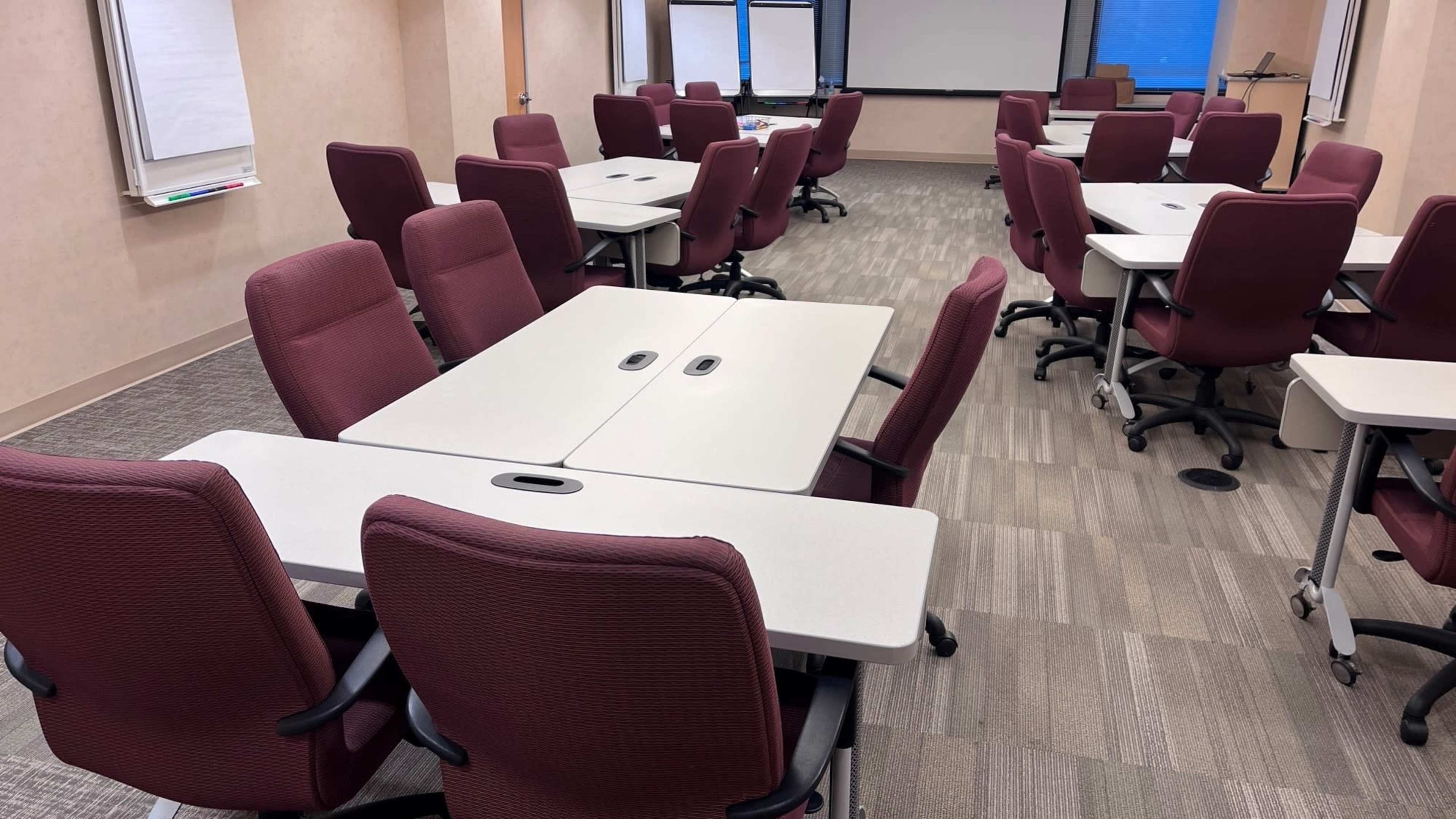 The image shows a classroom or meeting room arranged with multiple tables and chairs, all in neat rows, with large windows providing natural light.