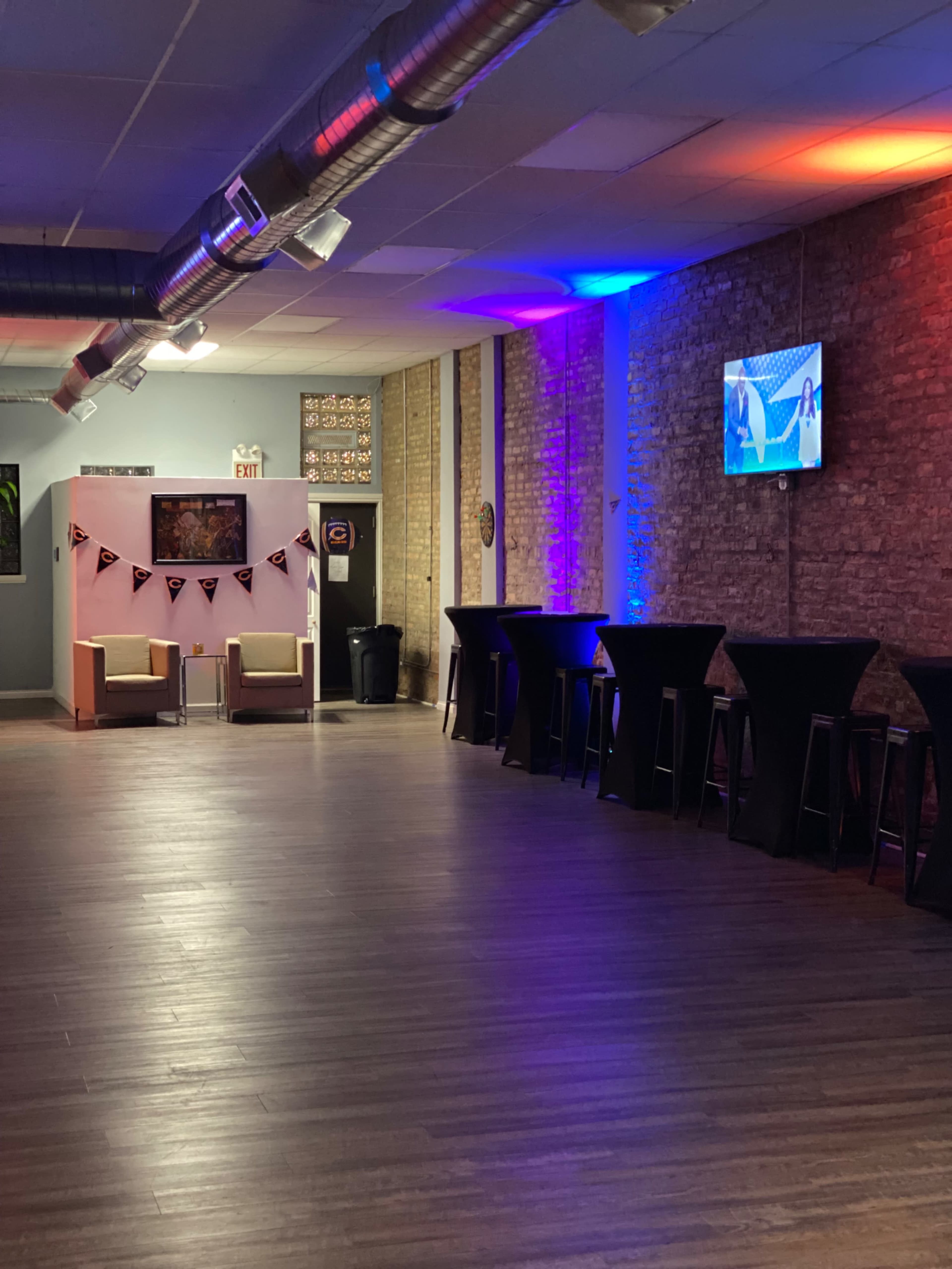 The image shows a spacious, dimly lit room featuring several tall tables, two seated areas, and a wall-mounted television displaying a scene.