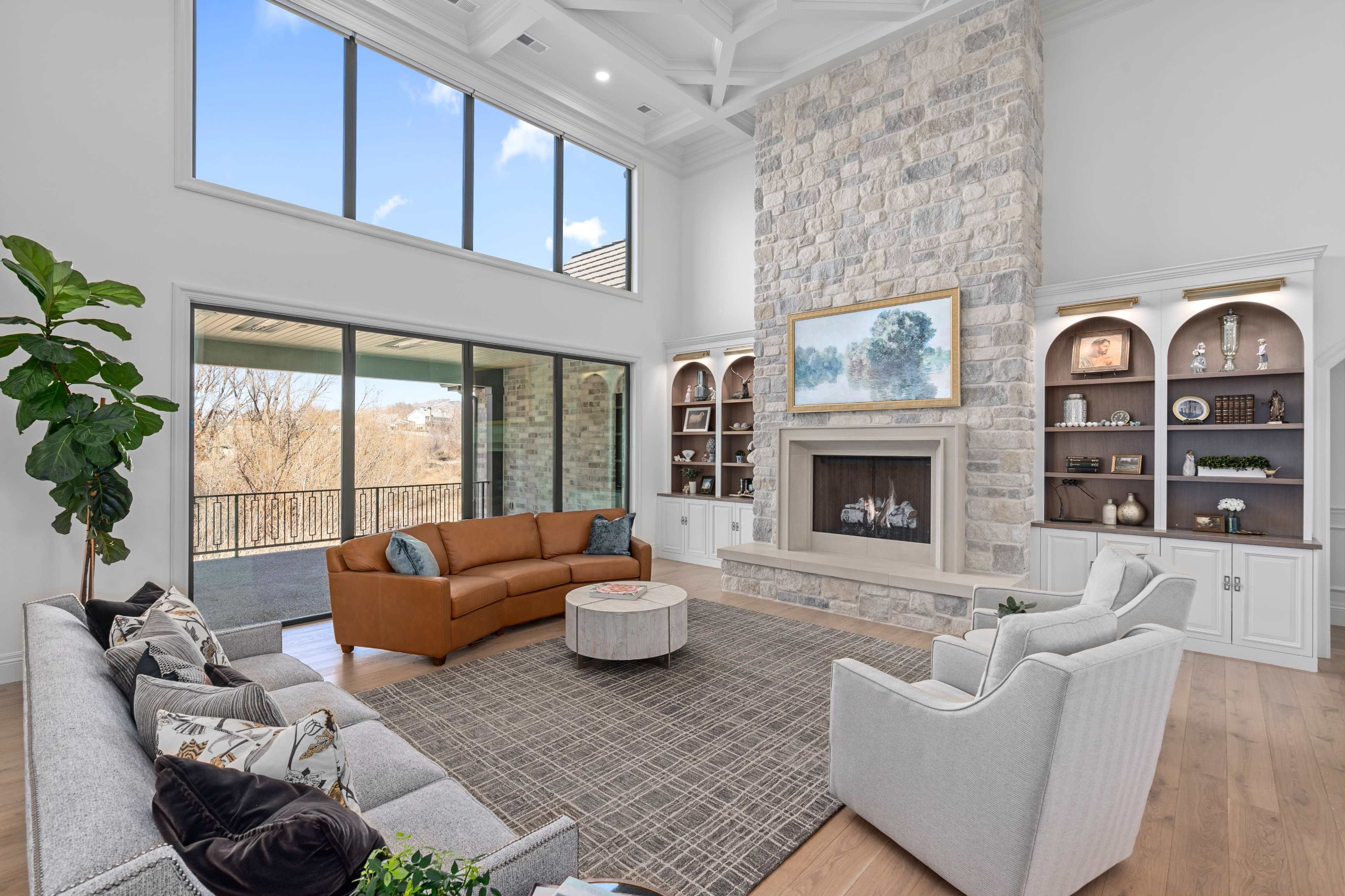 A spacious living room features a large stone fireplace, large windows, and several seating arrangements, including a brown leather sofa and light-colored armchairs.