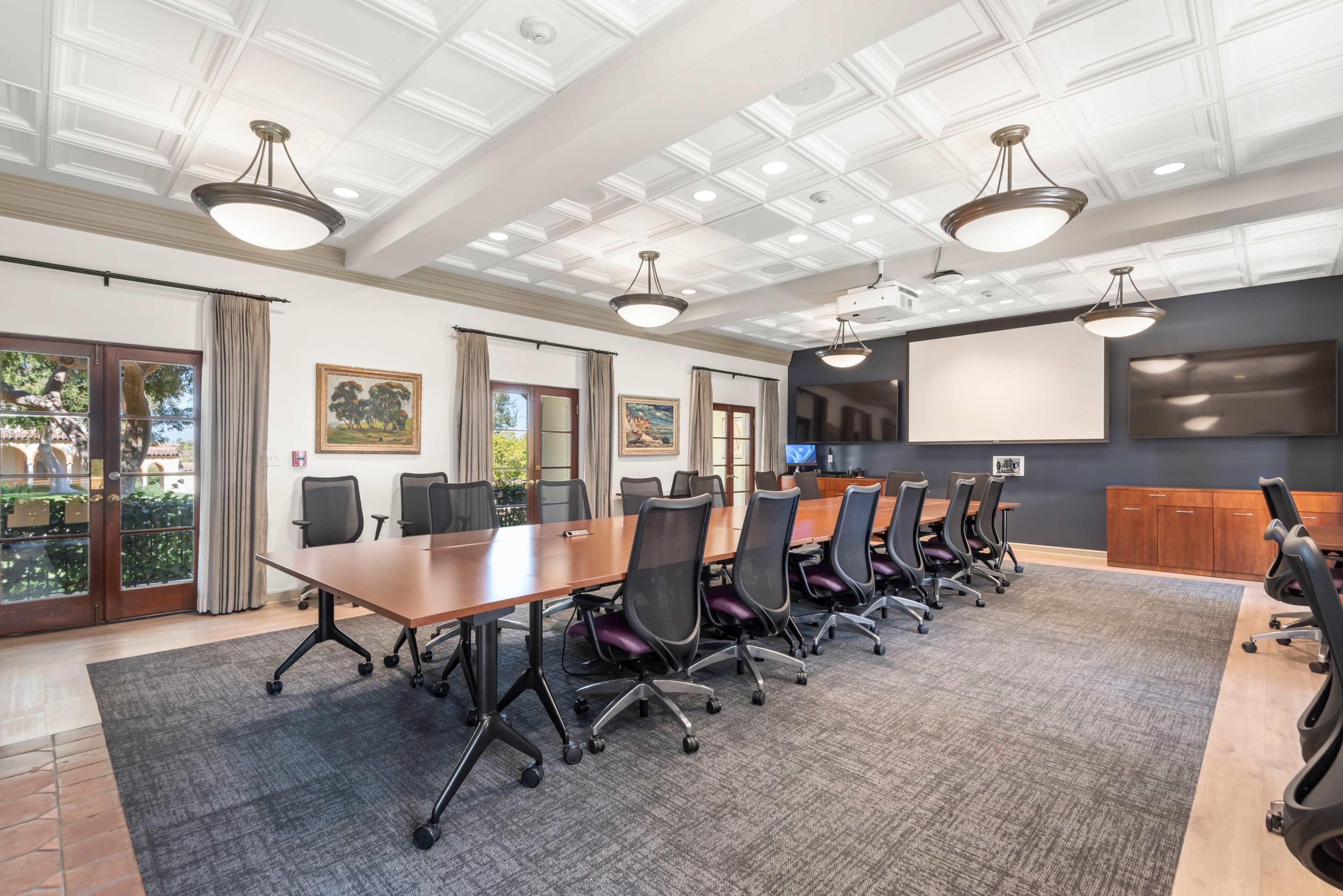 A modern conference room features a long table surrounded by ergonomic chairs, with a large screen mounted on the wall and windows that provide natural light.