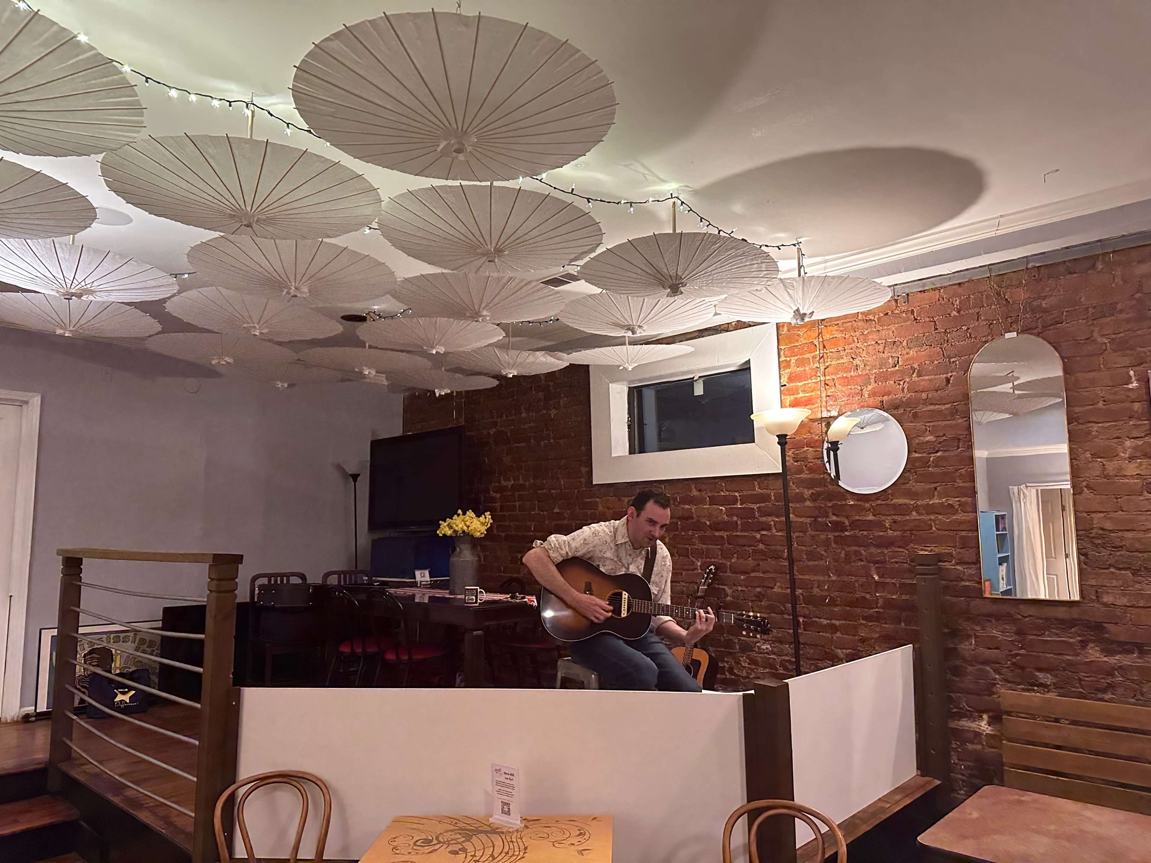 A musician plays guitar in a cozy cafe adorned with paper umbrellas on the ceiling and exposed brick walls.