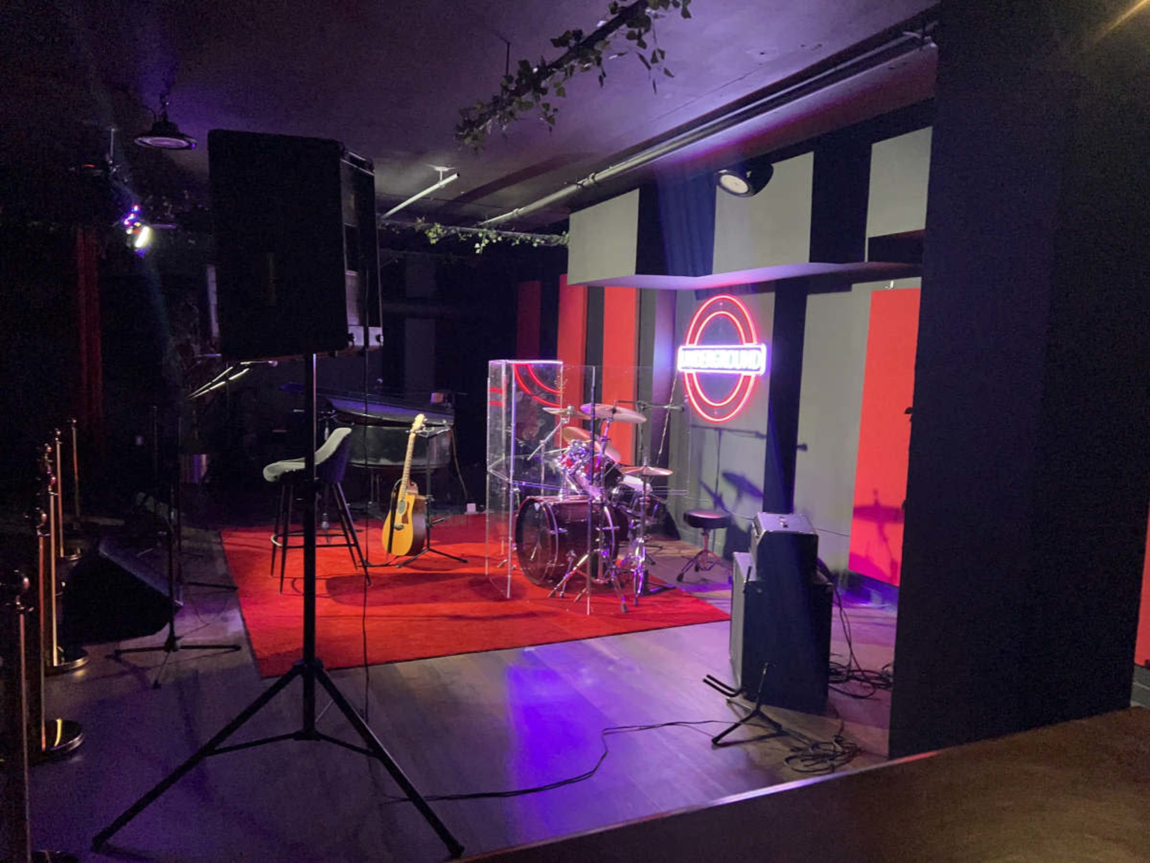 The image shows a music stage set up with a drum kit enclosed in a transparent soundproof chamber, flanked by a guitar and various audio equipment.