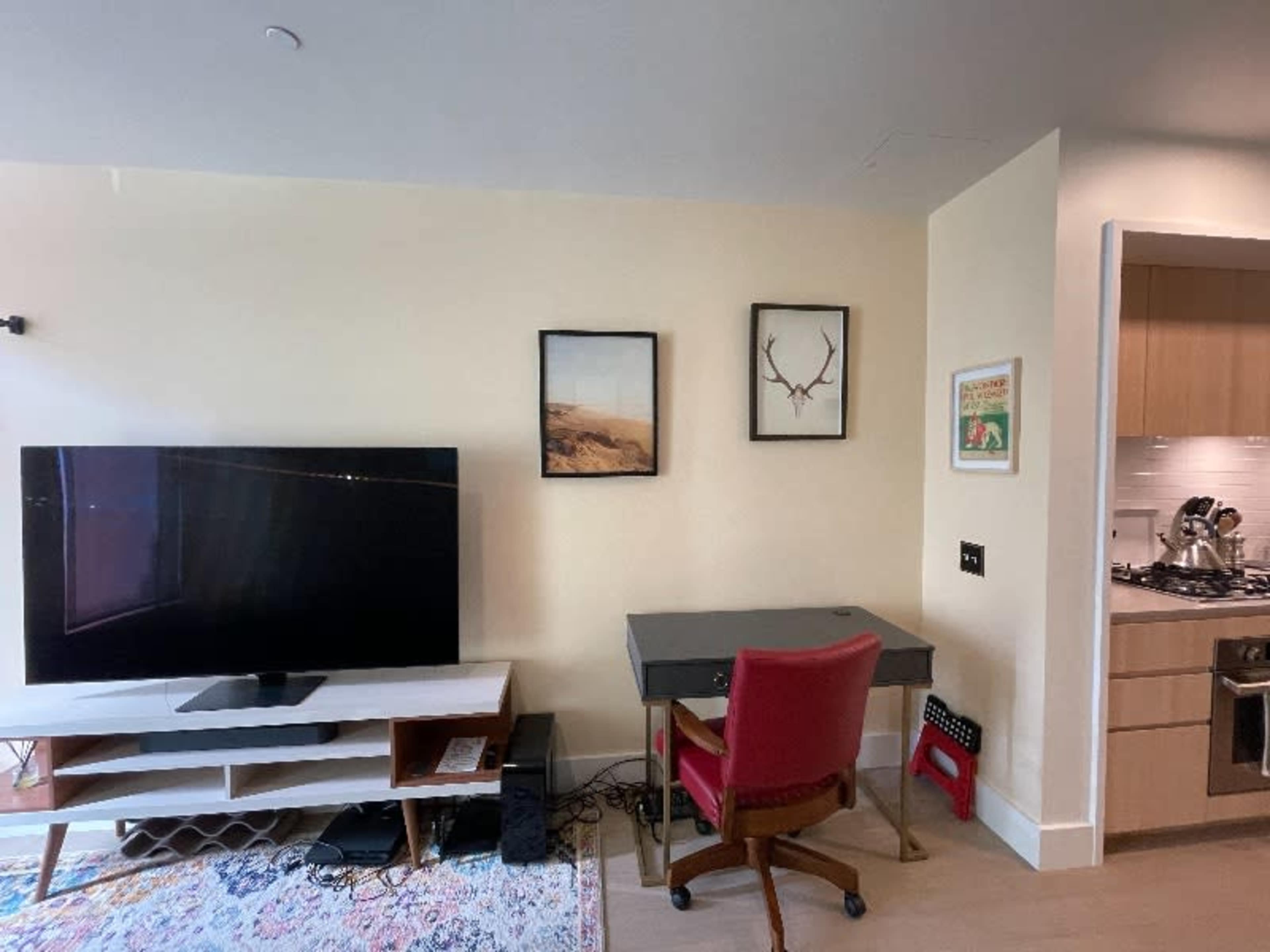 A living area featuring a large television mounted on the wall, a gray desk with a red chair, and framed artwork on the wall.
