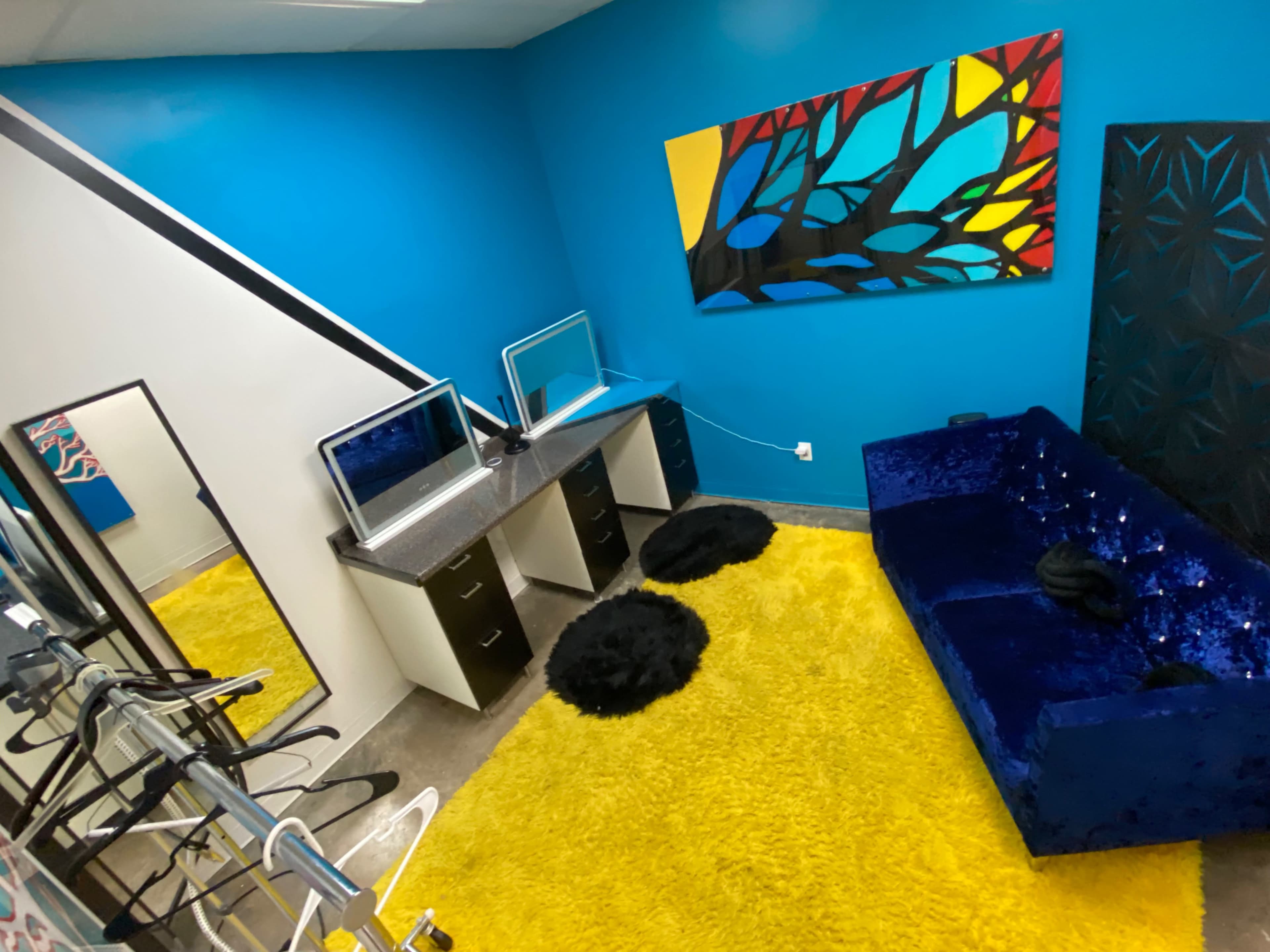 The image shows a brightly colored room with a blue wall, a yellow carpet, two mirrored vanity stations, and a blue couch, alongside a black textured panel and clothing racks.