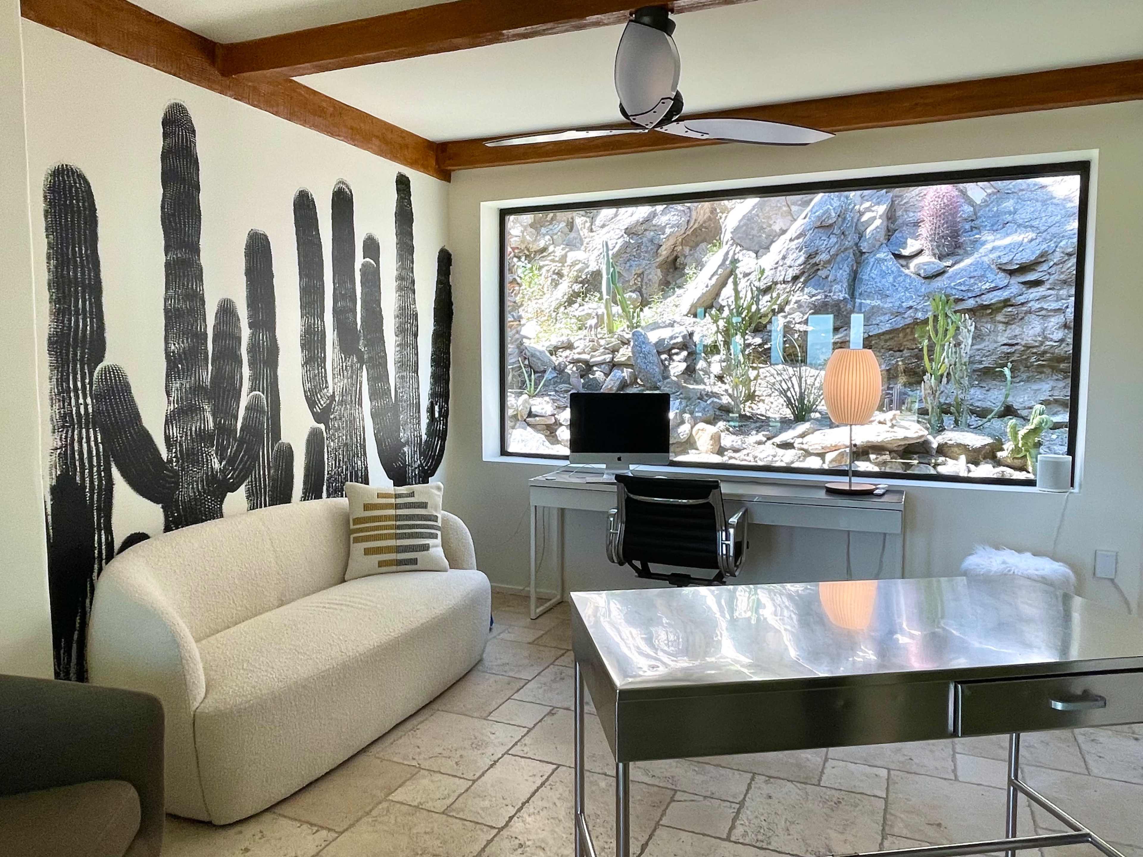 A modern workspace with a cacti-themed wall mural, a desk with a computer, and a large window overlooking a rocky garden with various plants.