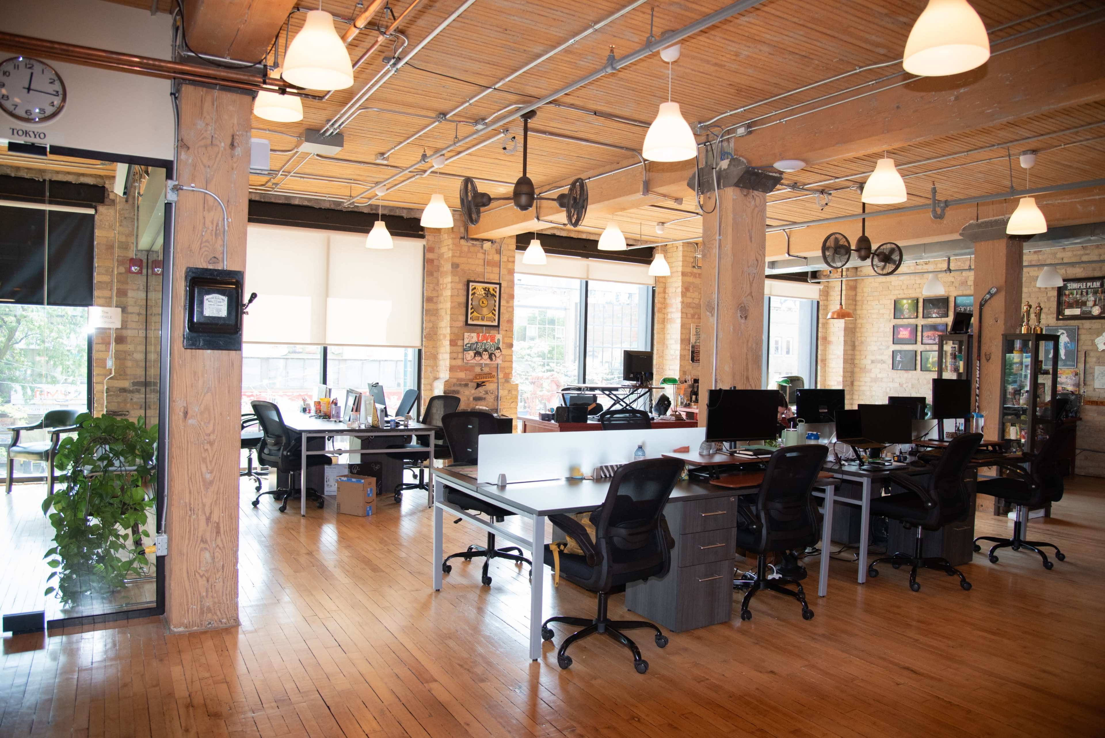 The image shows a modern office space featuring multiple desks with computers, wooden floors, exposed beams, and large windows allowing natural light.