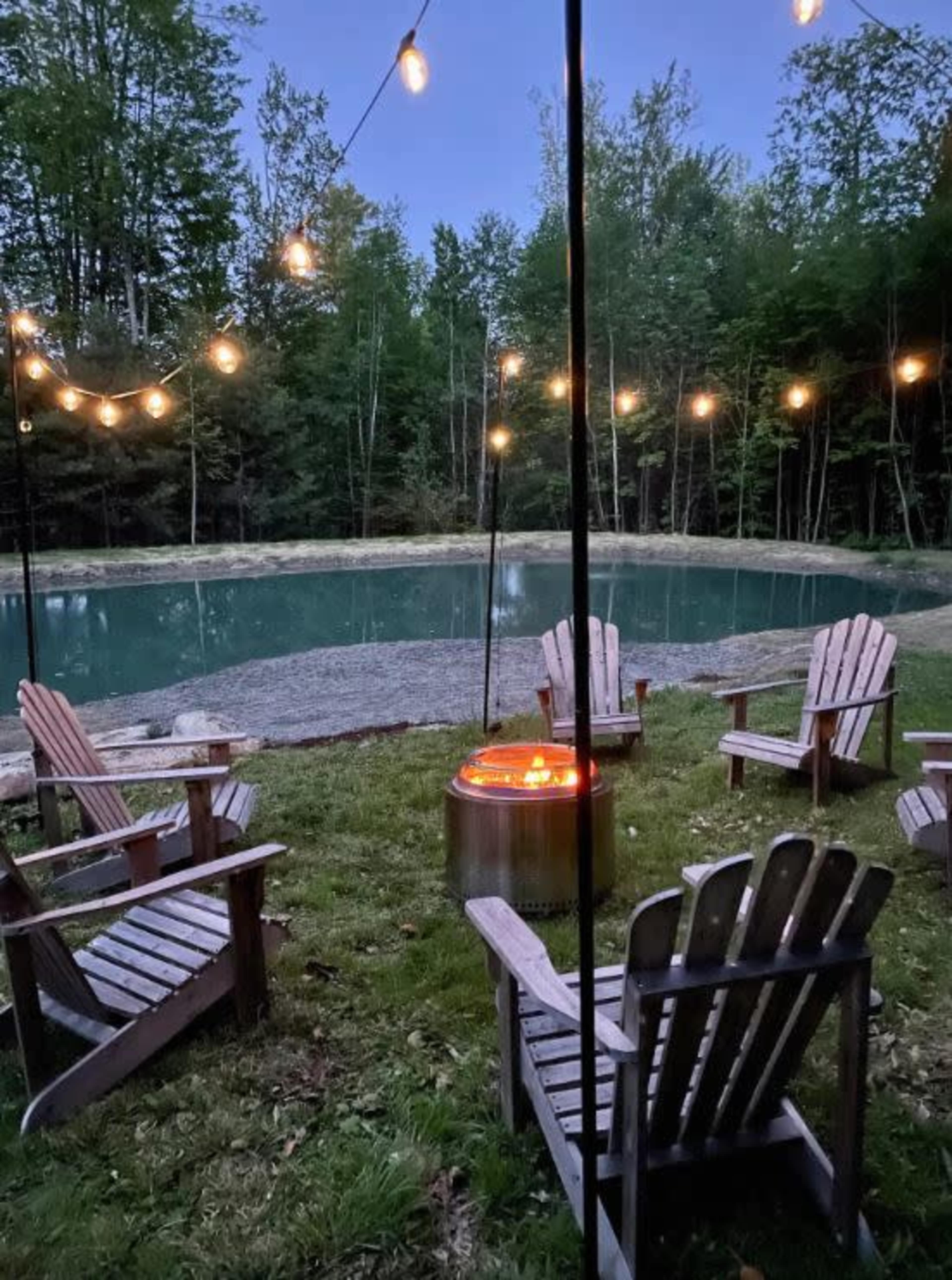 A circle of Adirondack chairs surrounds a fire pit near a pond, illuminated by string lights in a wooded area.