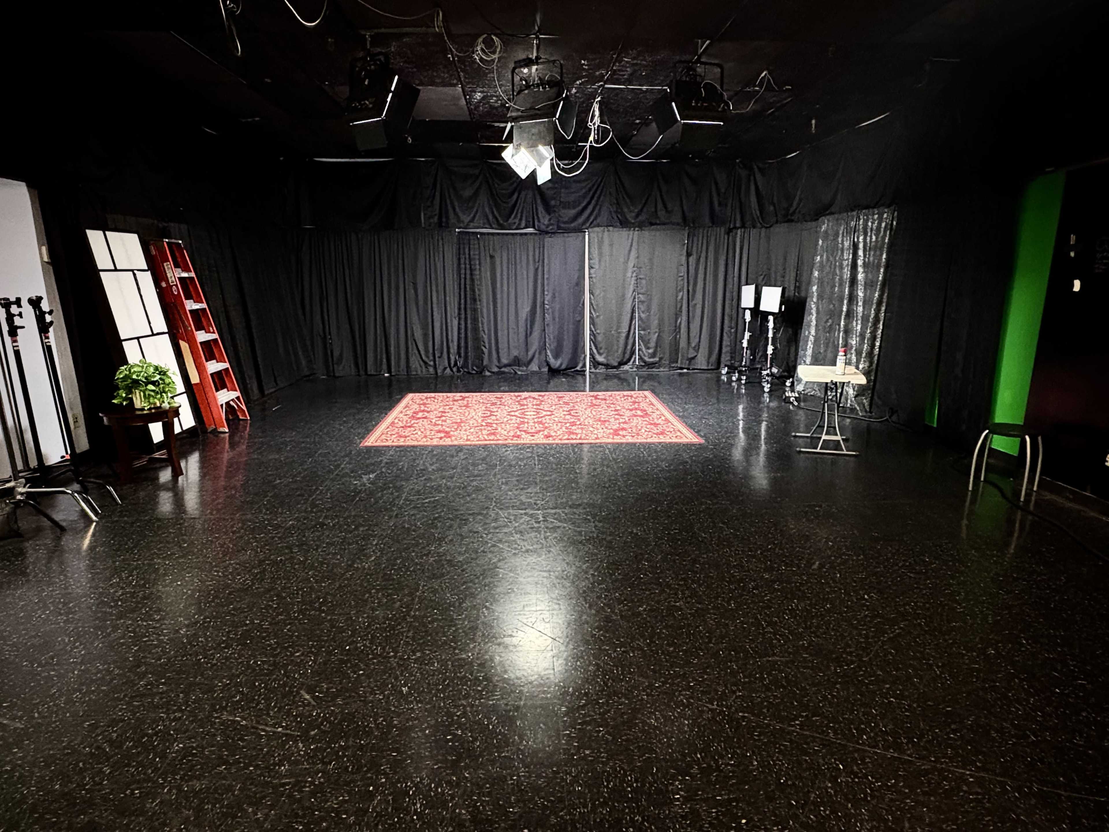 The image shows an empty studio space with black walls, a red rug in the center, and various lighting equipment positioned around the room.