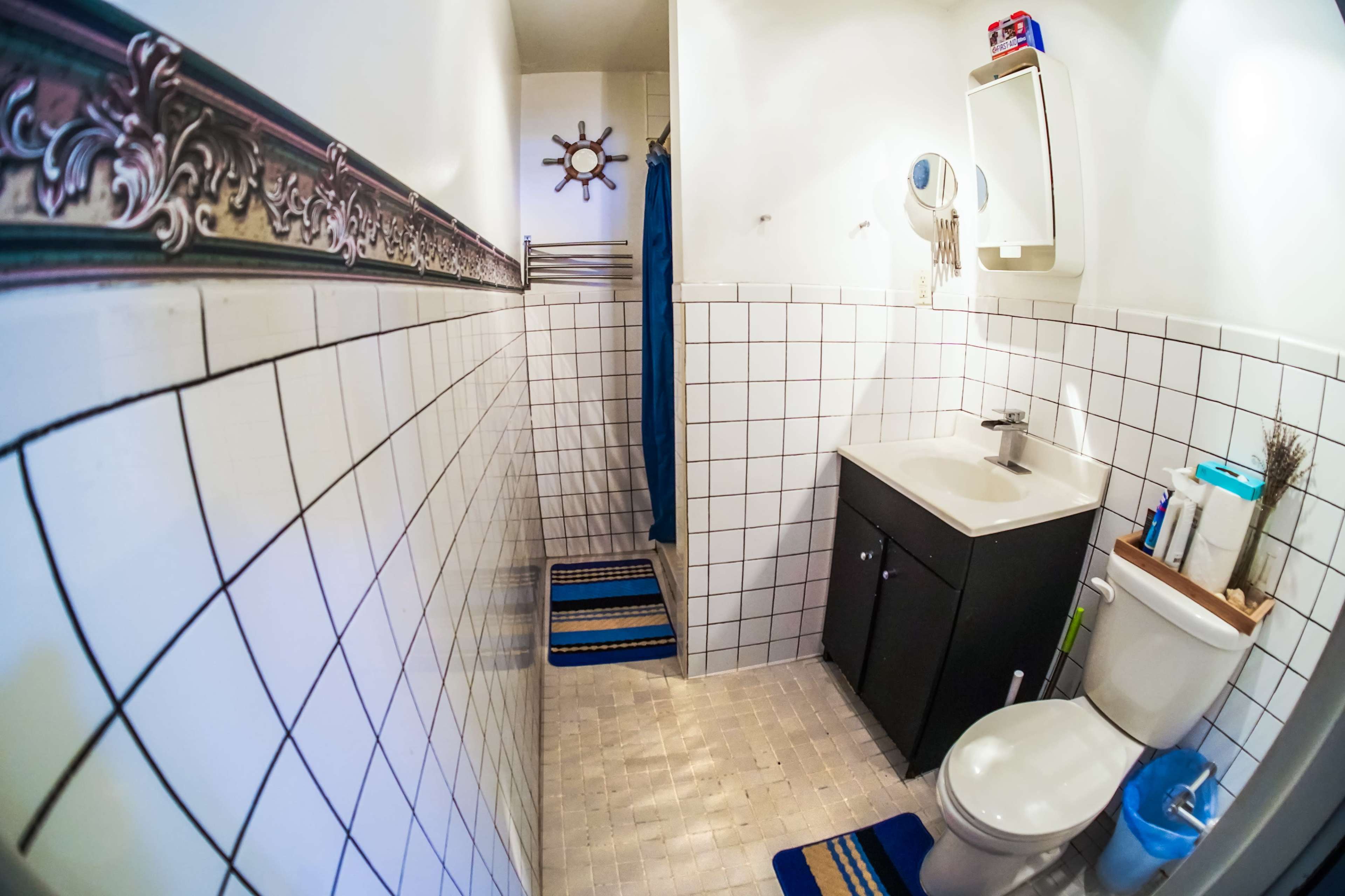 The image depicts a small bathroom featuring white tiled walls, a shower area with a blue curtain, a sink with a mirror, and a toilet.