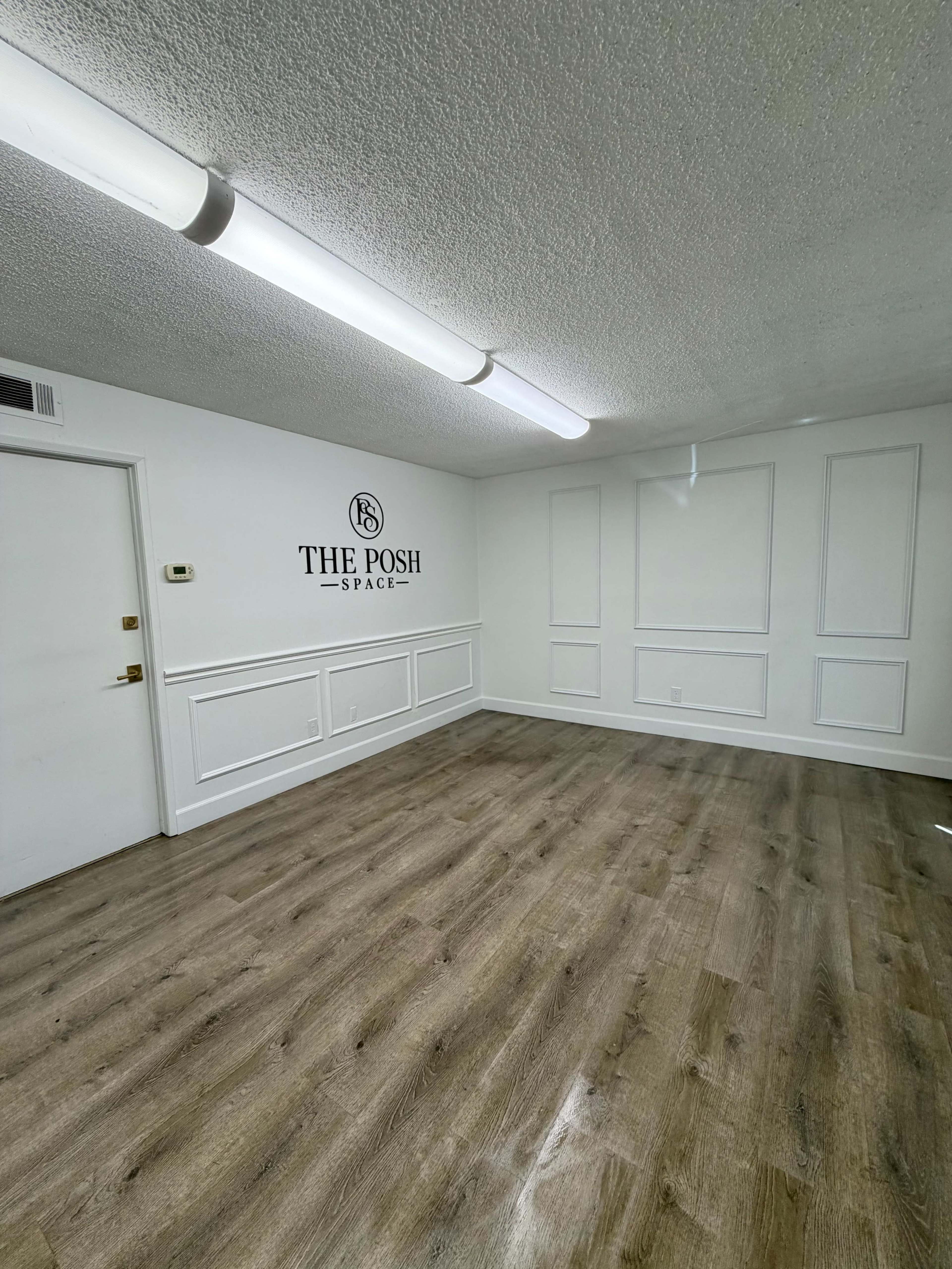 The image shows a spacious, empty room with light wood flooring and a white wall featuring a logo that reads "THE POSH SPACE."