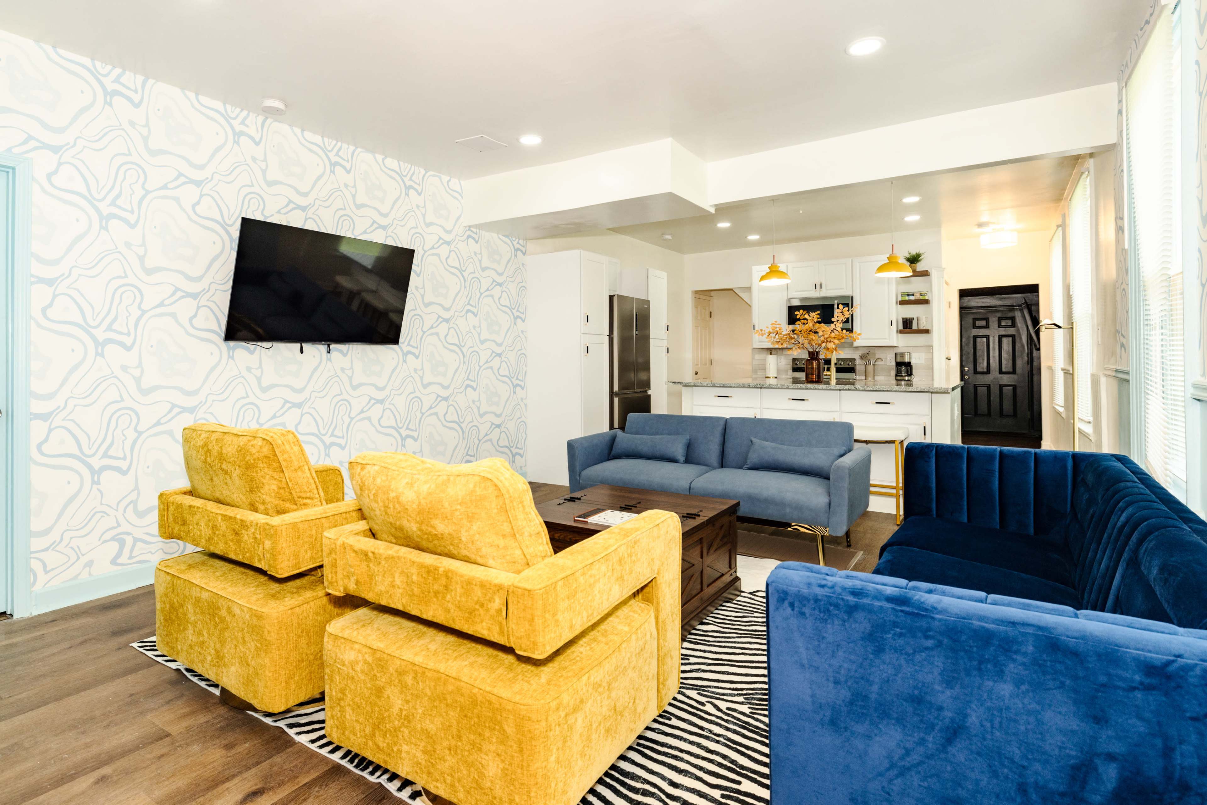 A modern living room with blue and yellow upholstered sofas, a wooden coffee table, and a patterned wallpaper, leading into a kitchen area.