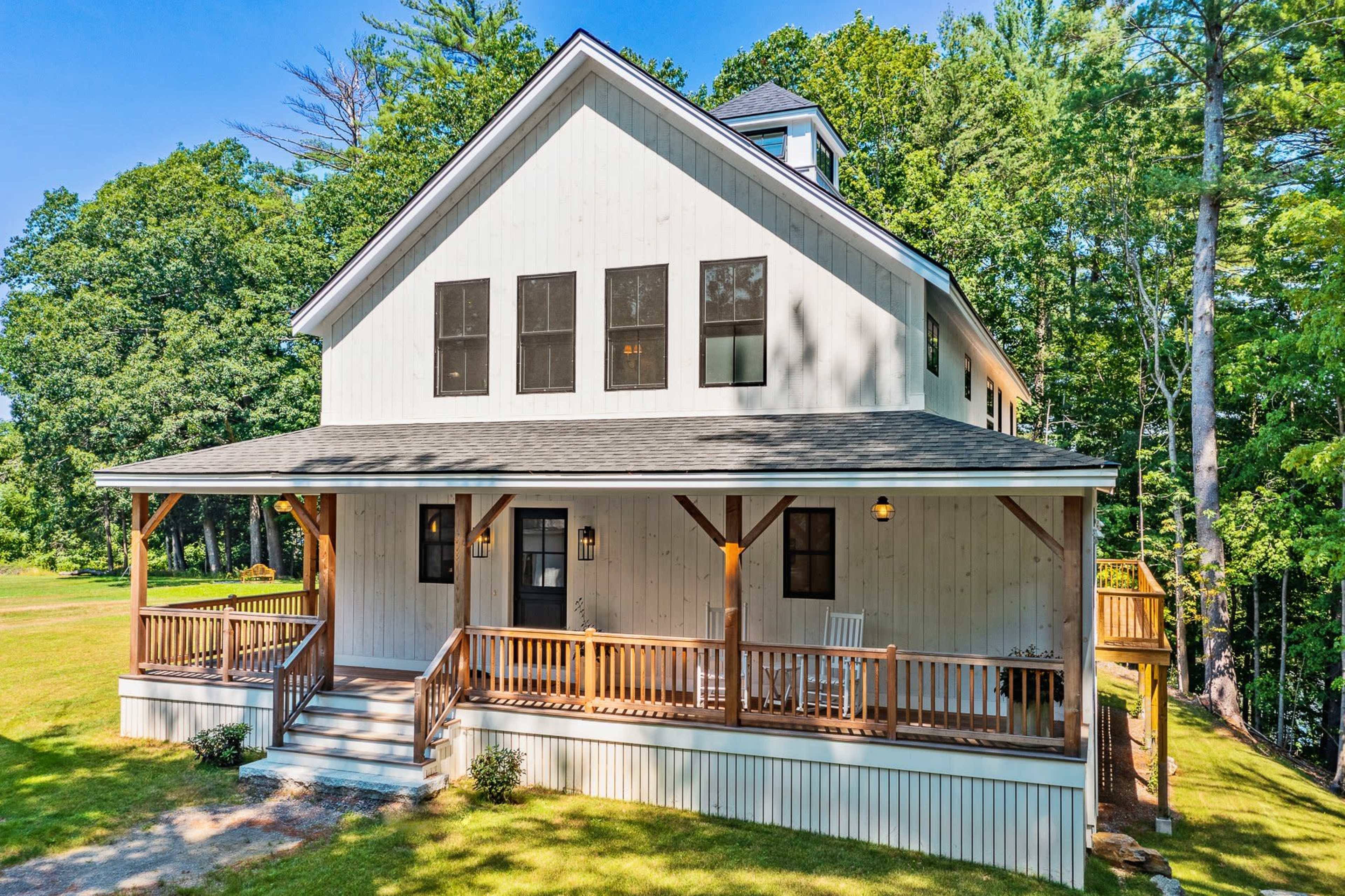 A two-story, modern-style house with a large front porch and a landscaped yard is surrounded by trees.