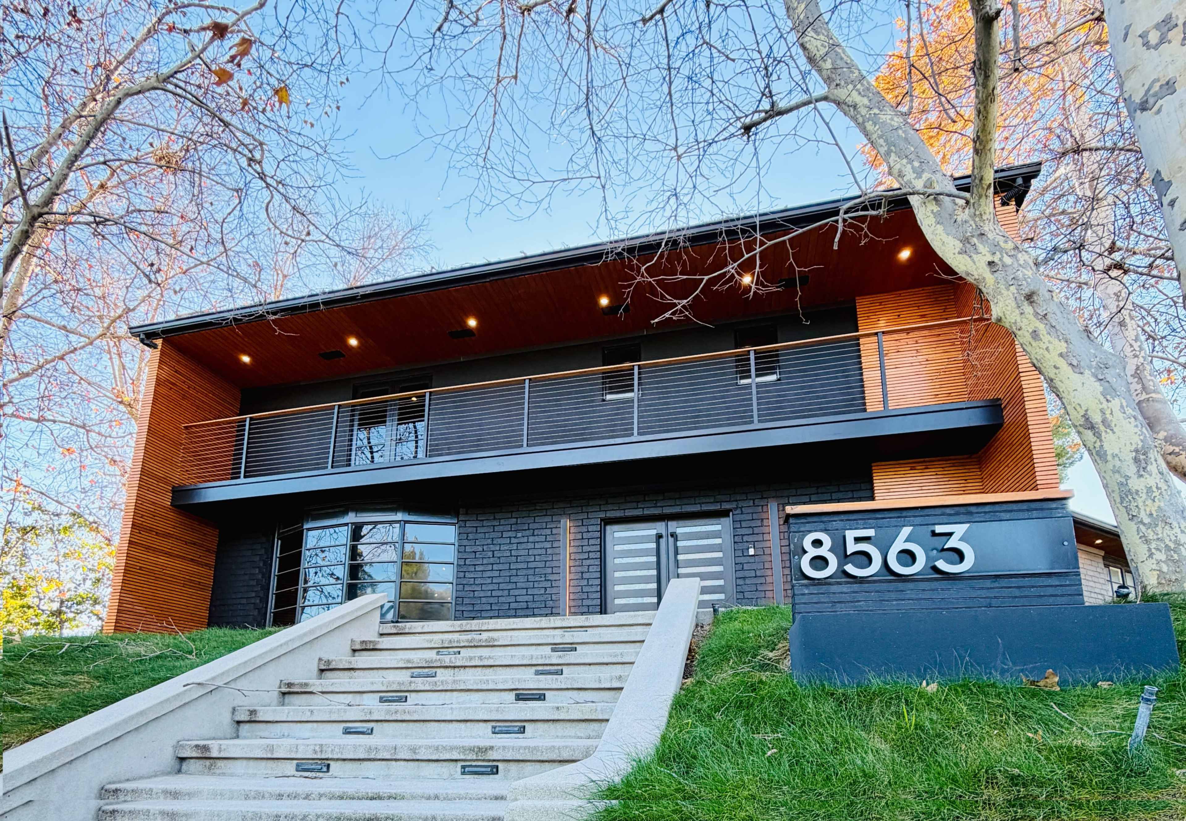 The image shows a modern two-story house with a mix of black and wooden exterior, featuring a prominent address sign and a set of stairs leading to the entrance.