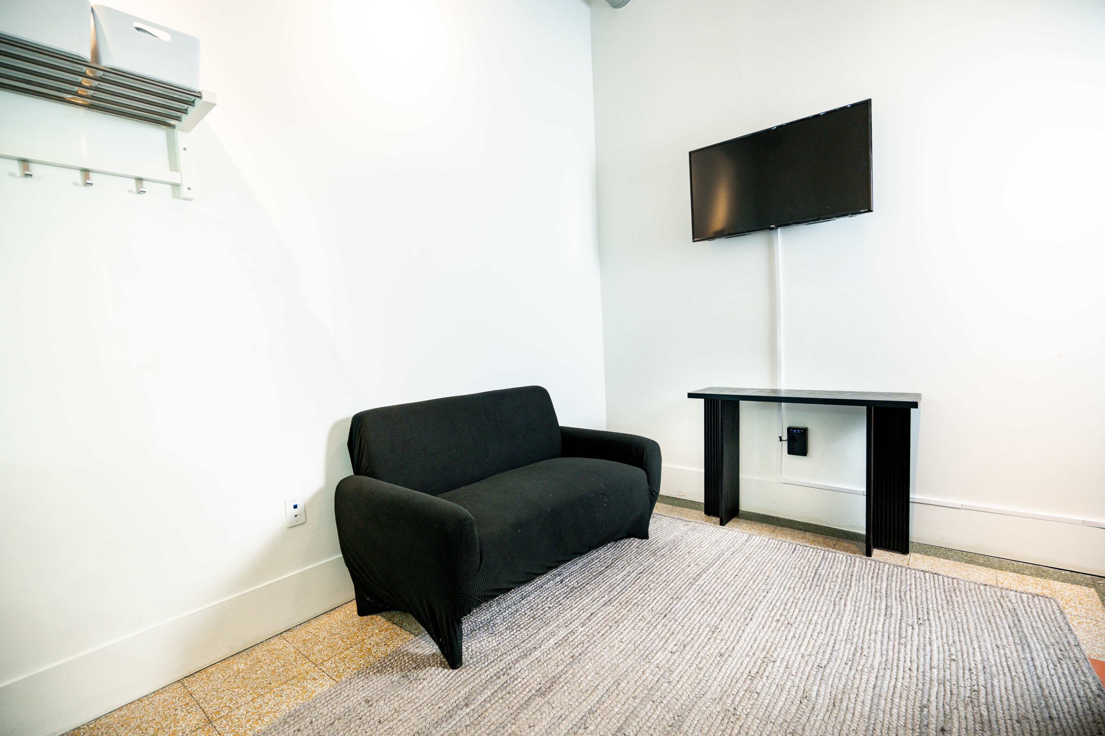 A simple room features a black sofa, a flat-screen TV mounted on the wall, and a small table on a rug.