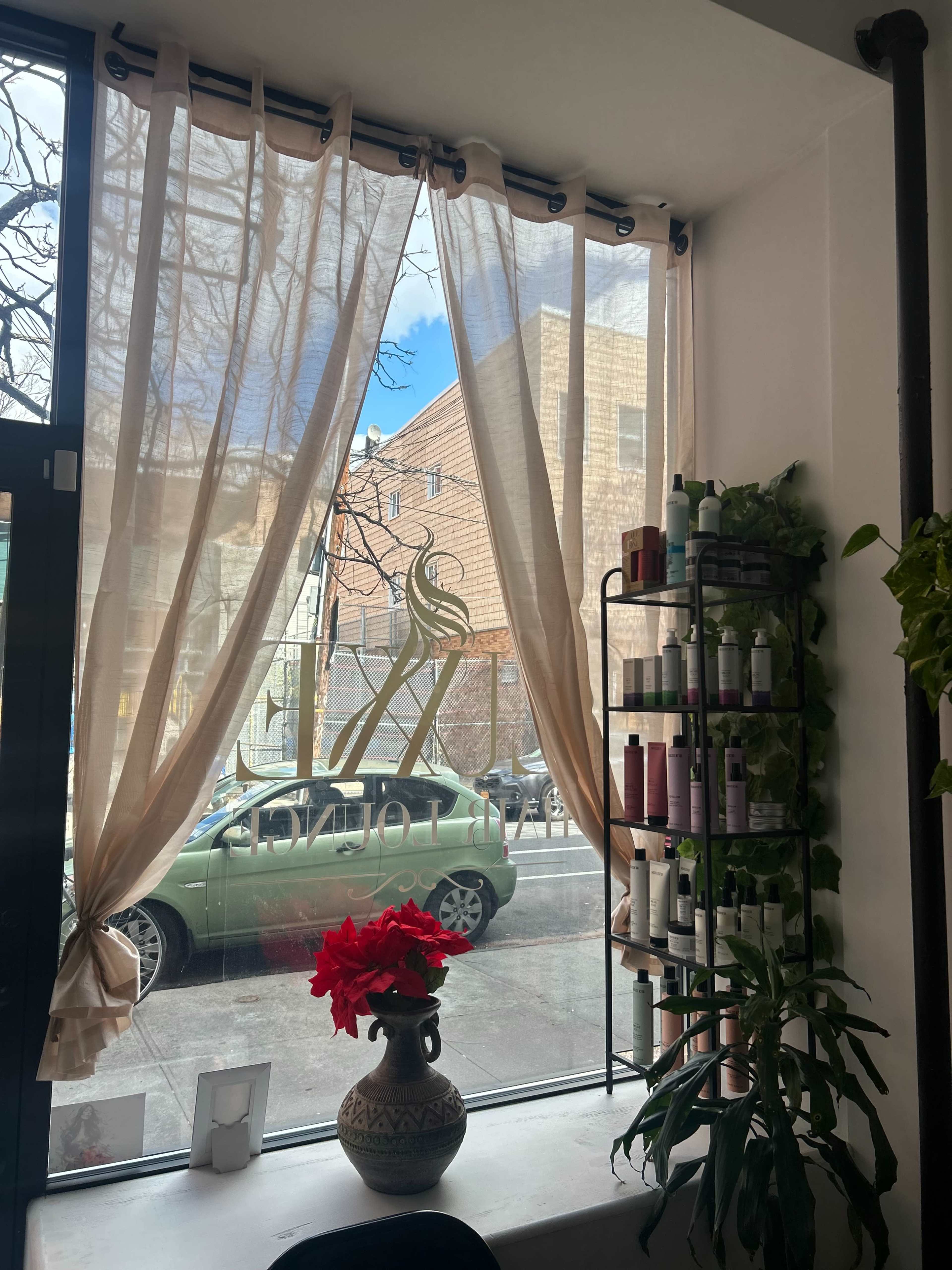 The image shows a salon interior with sheer curtains, a display of hair products on a shelf, and a view of a green car parked outside through the window.