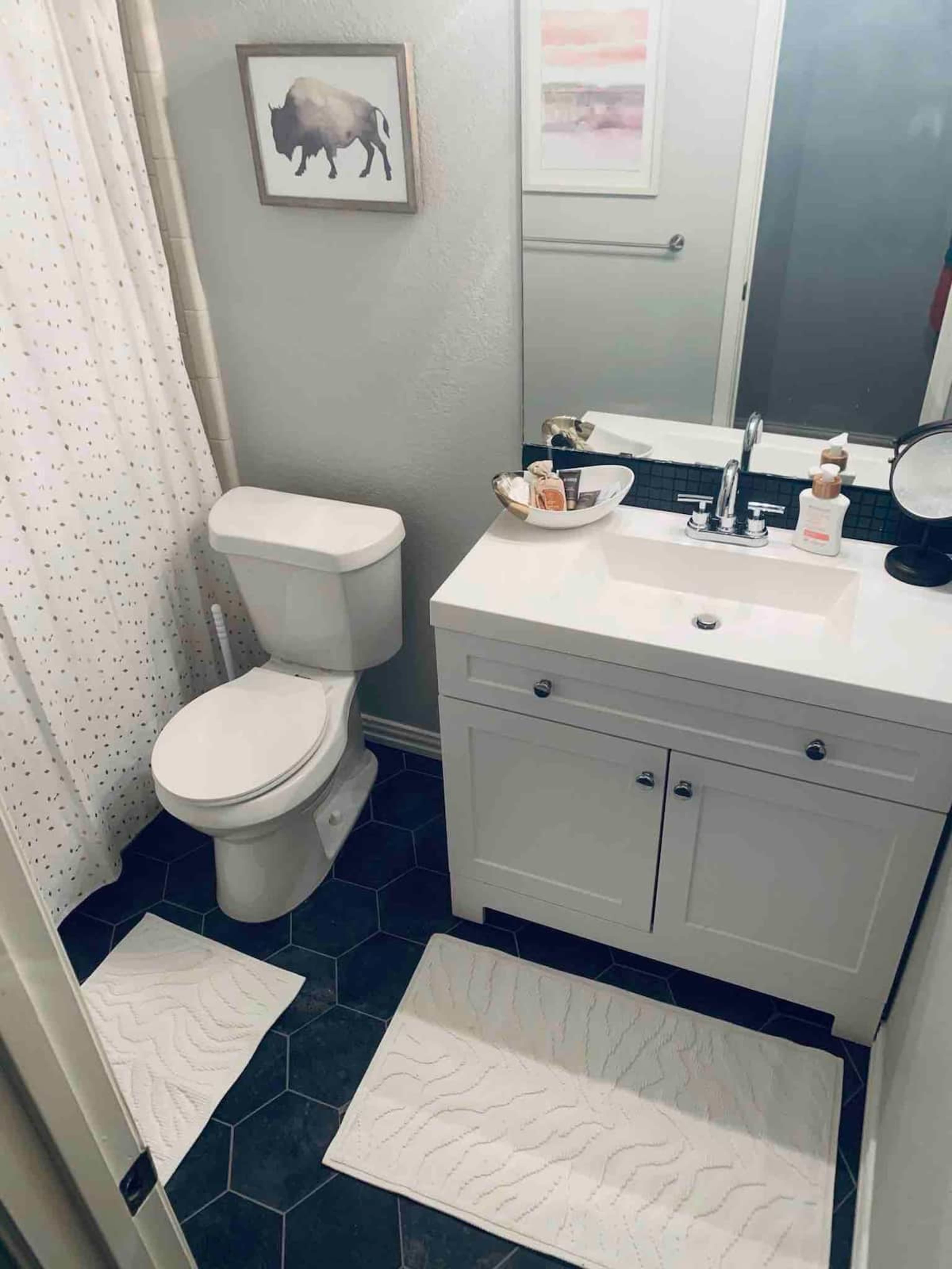 A small bathroom featuring a white toilet, a sink with a mirror above, and two textured bath mats on a hexagonal tiled floor.