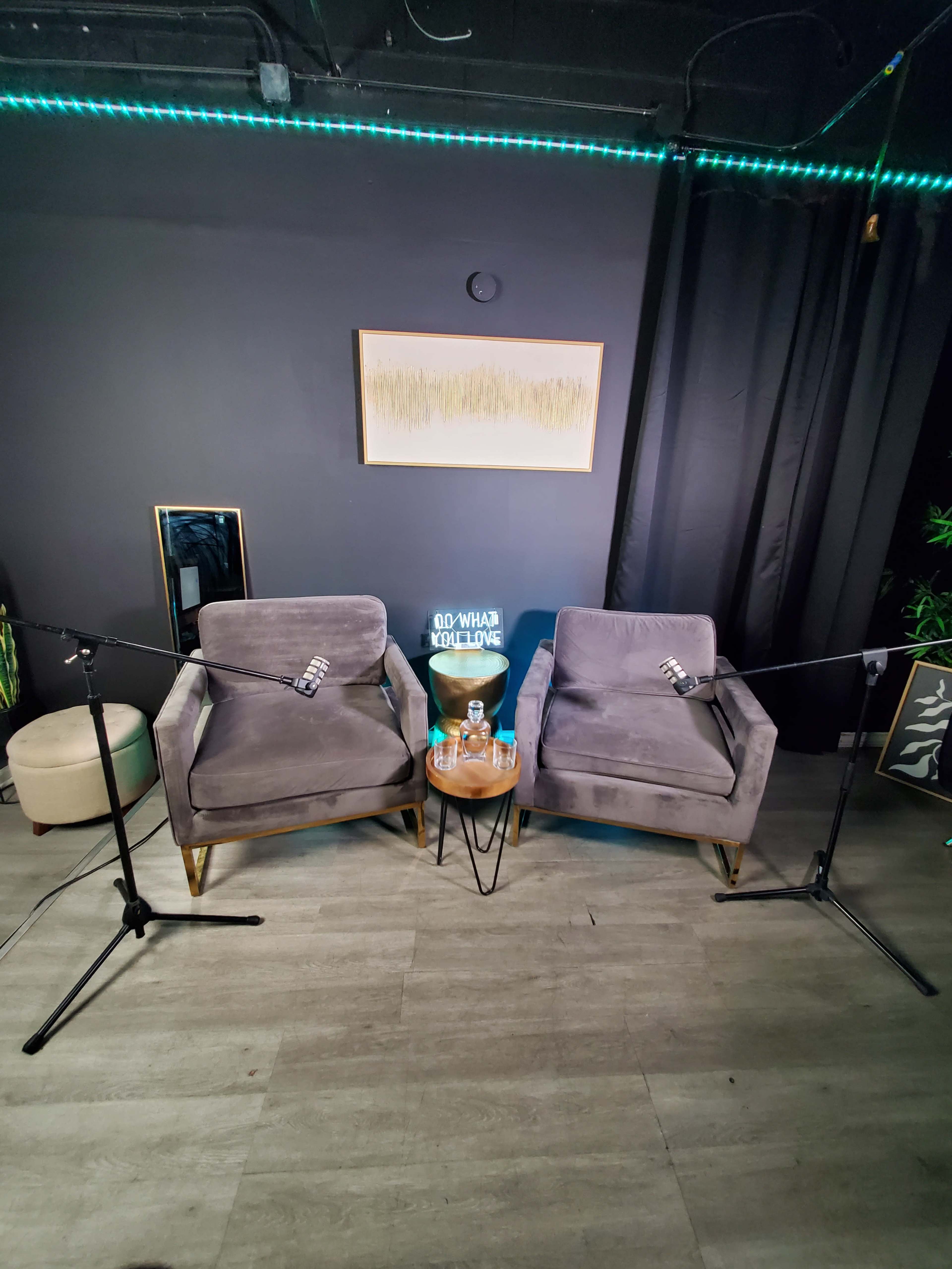 The image shows two gray chairs positioned opposite each other with microphones on stands and a small table between them, set in a room with dark walls and soft lighting.